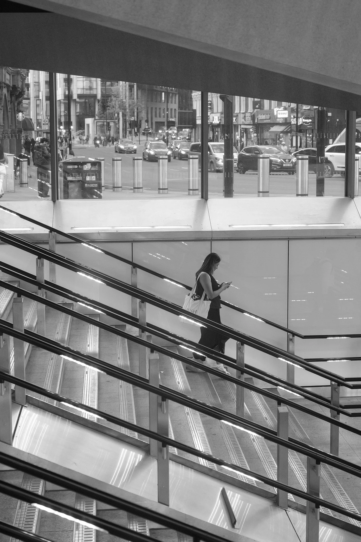 Une personne marche dans une station de métro en regardant son téléphone, vue depuis une rampe d'escalier. Un décor urbain visible par une grande fenêtre, avec des voitures, des piétons et des magasins.