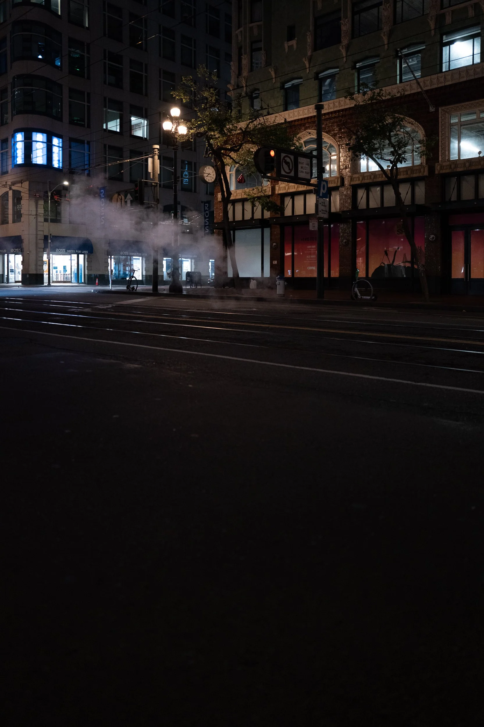 Vue nocturne d'une rue urbaine avec des bâtiments modernes, des arbres, de la vapeur ou de la fumée, un panneau de signalisation, une horloge, des lampadaires allumés et un vélo abandonné.