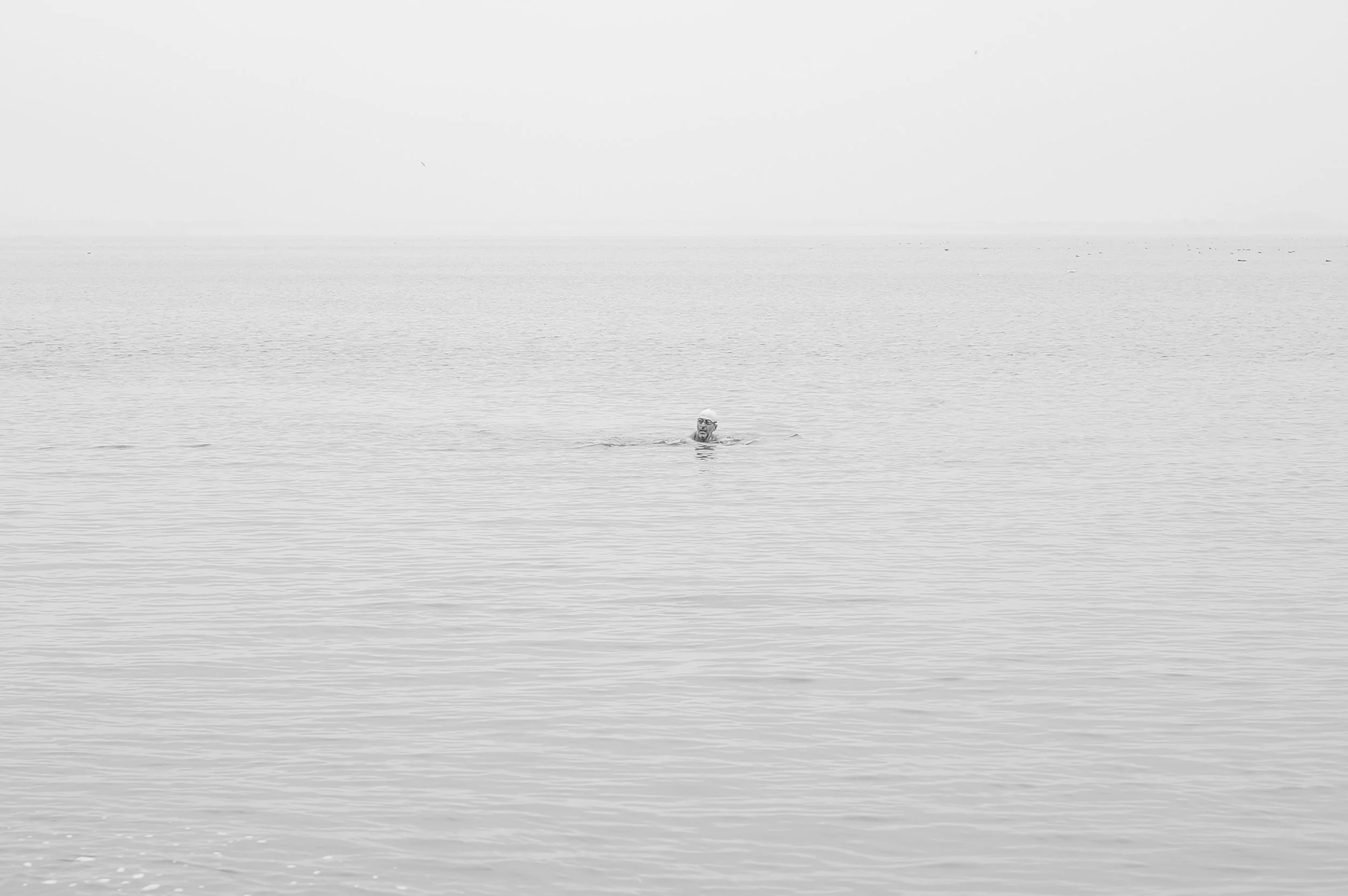 Une personne nage dans la mer avec une surface calme et un ciel nuageux, en noir et blanc.