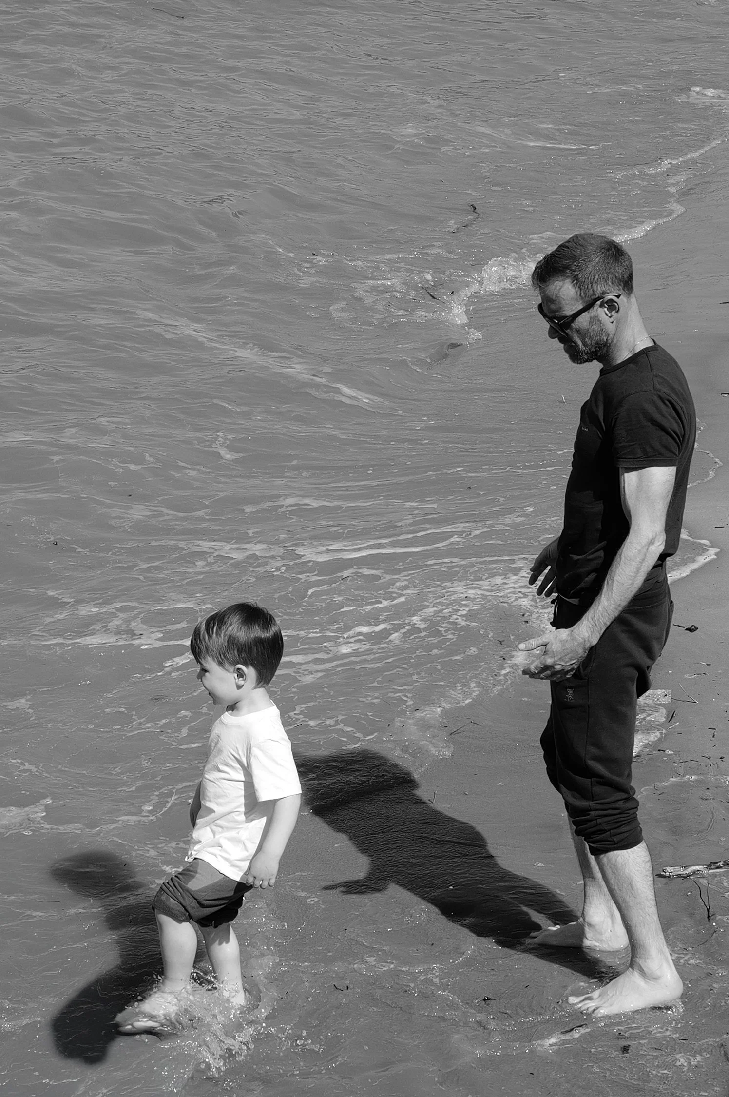 Un homme et deux enfants marchent pieds nus dans l'eau de la mer sur la plage, le paysage est en noir et blanc.