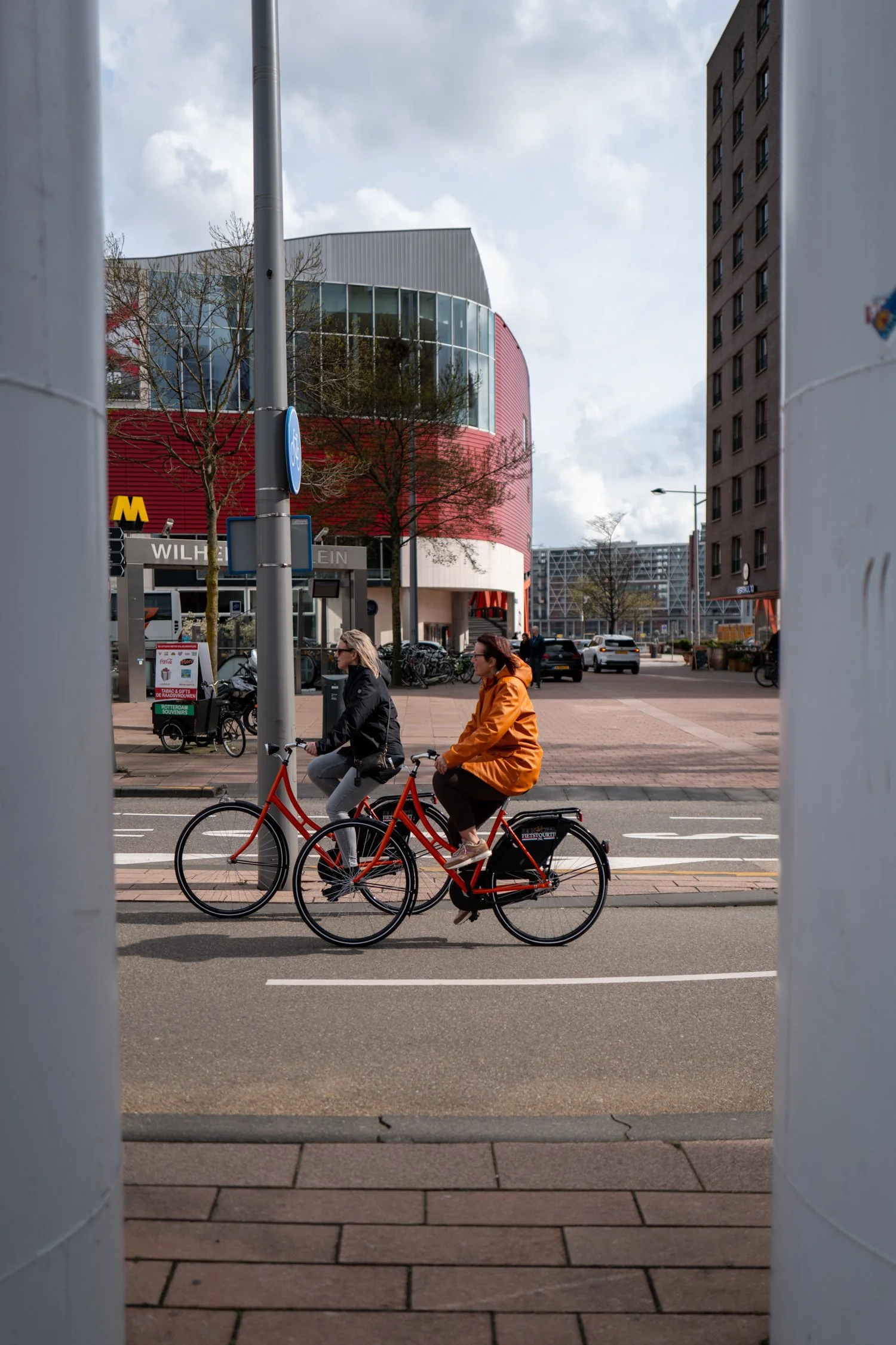 Deux personnes font du vélo sur une rue urbaine, en traversant entre deux barres blanches. Au fond, un bâtiment moderne avec des fenêtres en verre, des arbres et une rue avec des voitures.