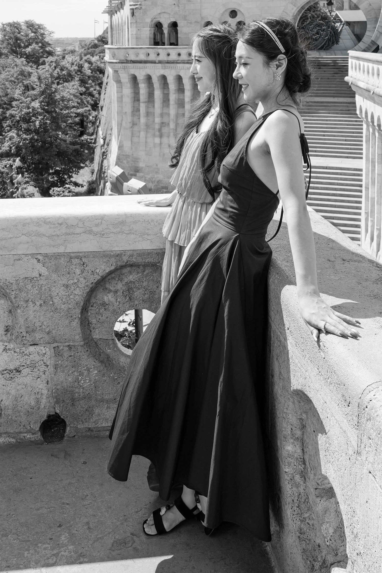 Deux femmes en robes de soirée noires se tiennent côte à côte sur un balcon en pierre, regardant à l'horizon, avec une architecture ancienne en arrière-plan.