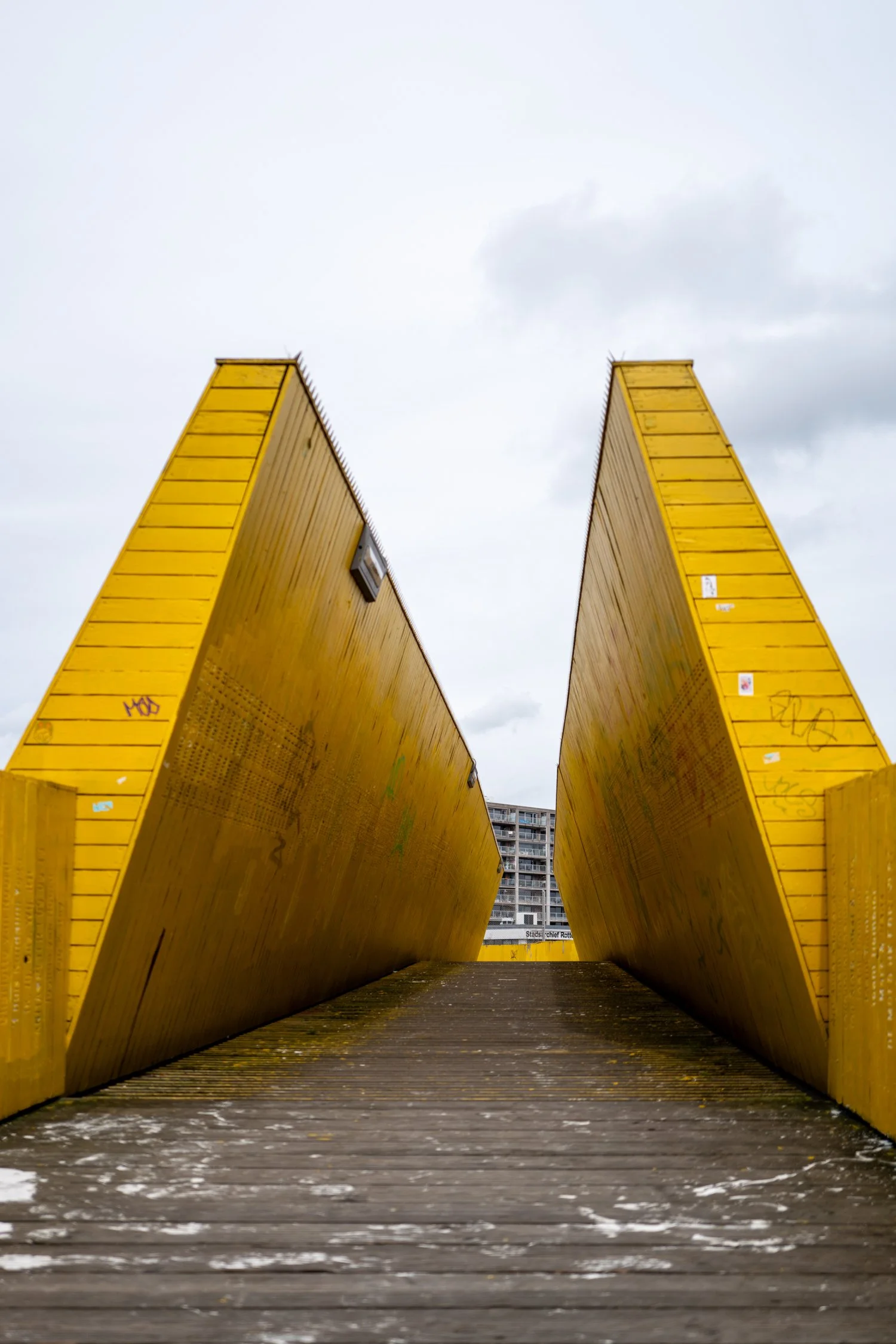 Vue d'une passerelle ou un pont piéton en bois avec deux structures jaunes en forme de triangles inversés, dans une zone urbaine, avec un bâtiment résidentiel en arrière-plan et un ciel nuageux.