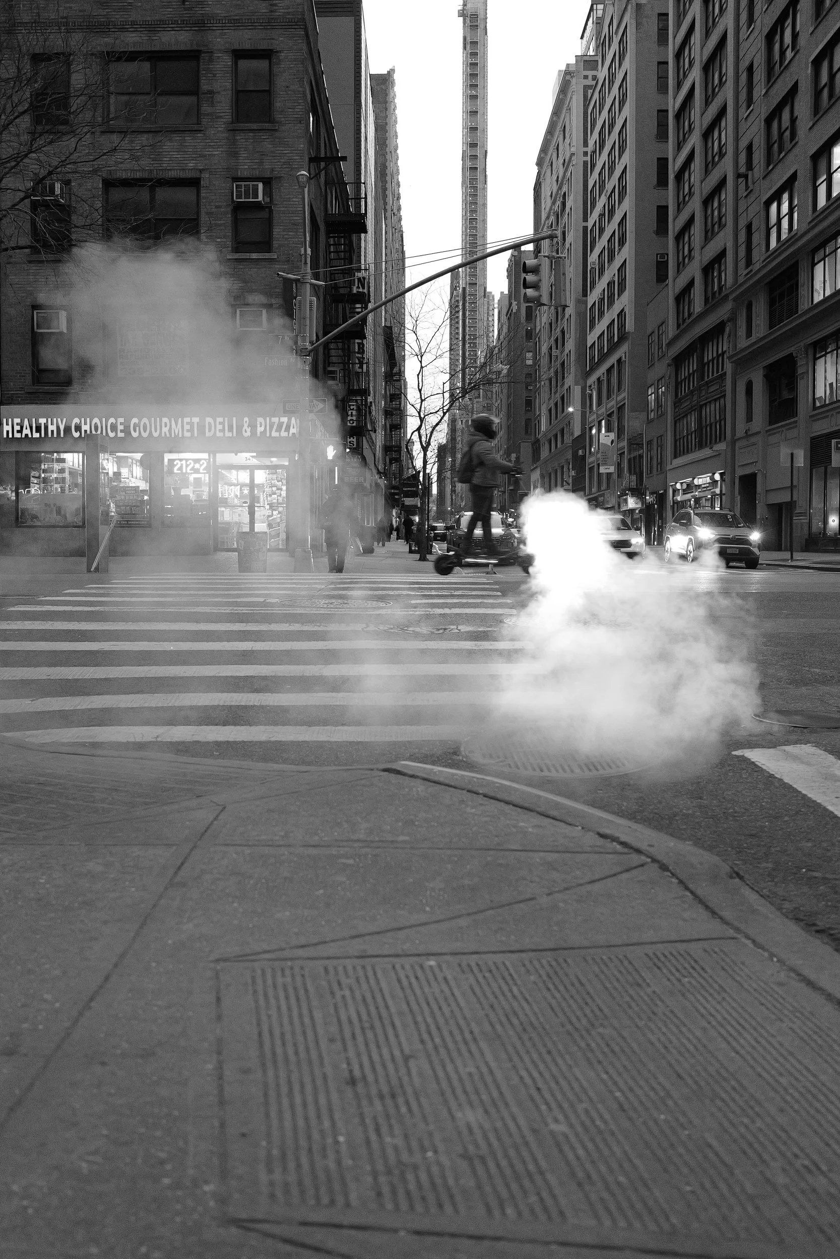 Une rue urbaine en noir et blanc avec des bâtiments, des voitures, une personne à skate électrique, de la vapeur sortant d'une bouche d'incinérateur, et un panneau pour une pizzeria.