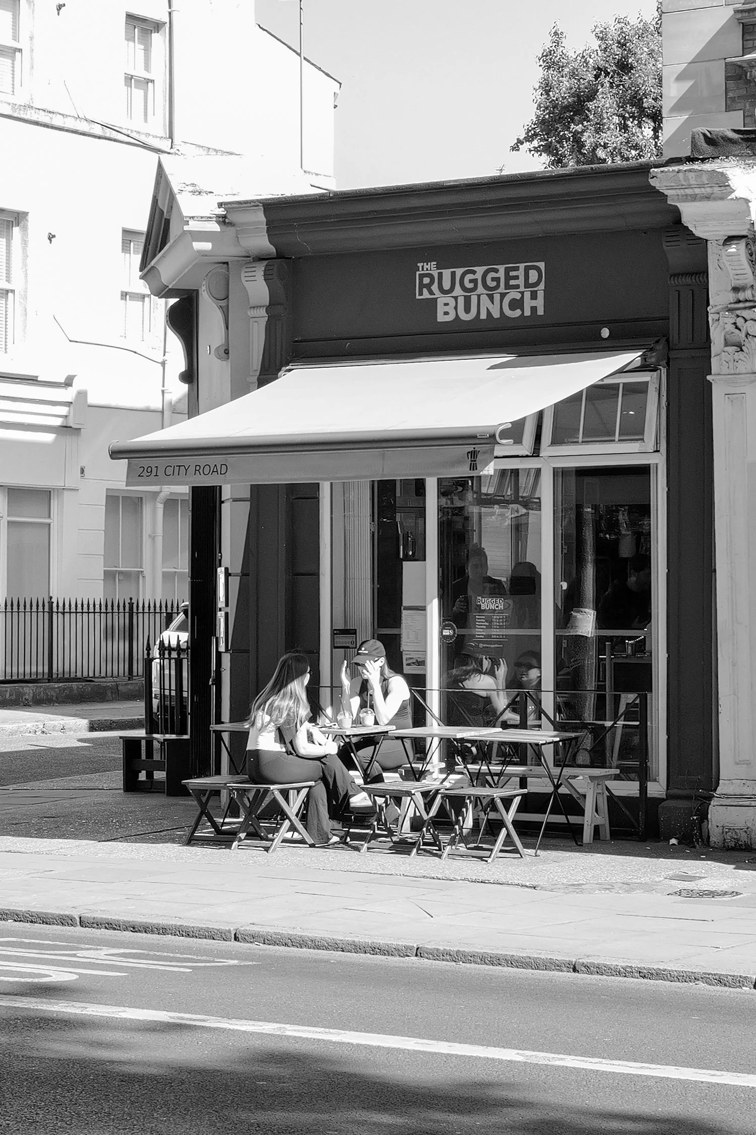 Une scène de rue avec une terrasse de café où deux femmes discutent assises à une table, avec un bâtiment en arrière-plan et un panneau indiquant l'adresse 291 City Road. L'enseigne au-dessus du café affiche 'The Rugged Bunch'.