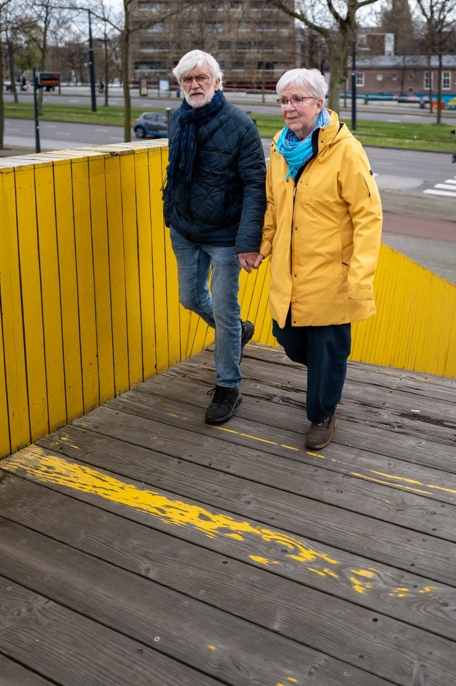 Un couple âgé marche main dans la main sur un pont en bois avec une rambarde jaune dans une ville. Il fait un temps frais et nuageux, avec des arbres sans feuilles en arrière-plan.
