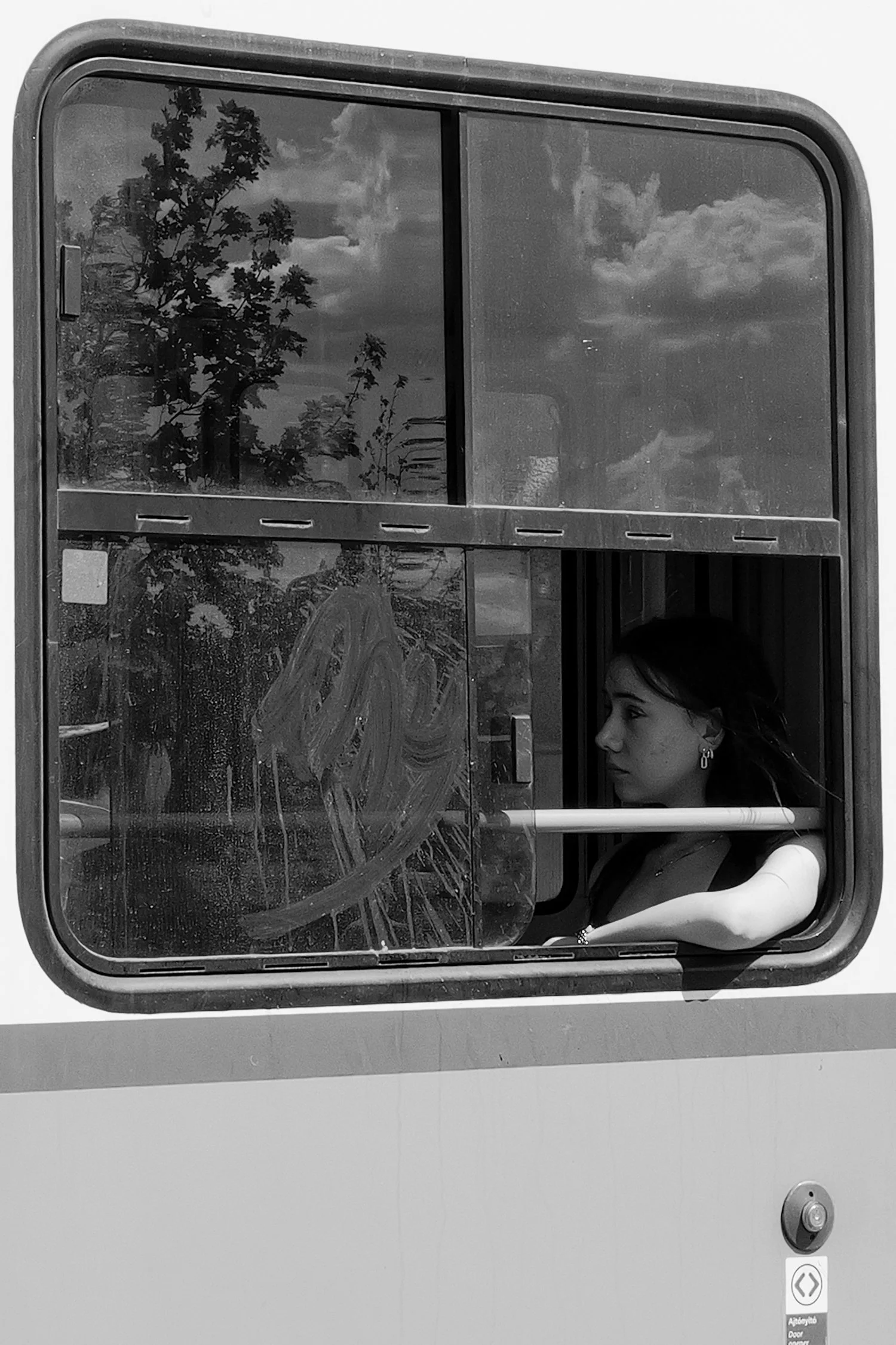 Une femme regarde par une fenêtre d'un bus ou d'un tramway, avec des arbres et un ciel nuageux visibles à travers le verre.