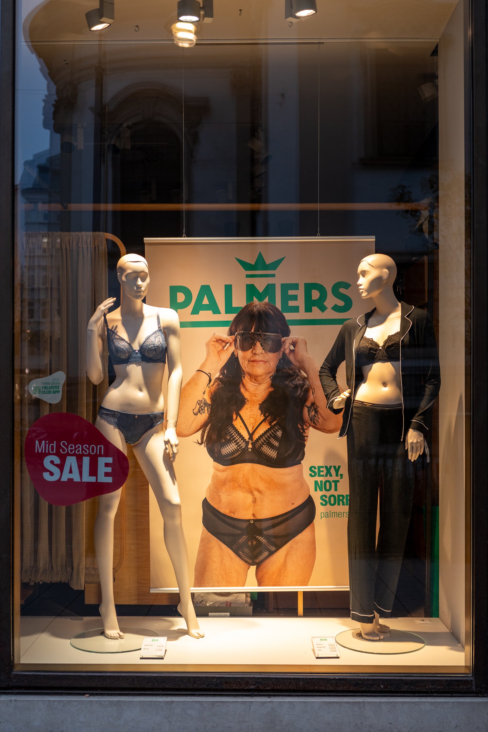 Vitrine d'un magasin de lingerie avec deux mannequins portant des sous-vêtements. Au centre, une grande affiche montrant une femme en lingerie avec le texte 'SEXY, NOT SORRY'. Un panneau annonce une 'Mid Season SALE'.