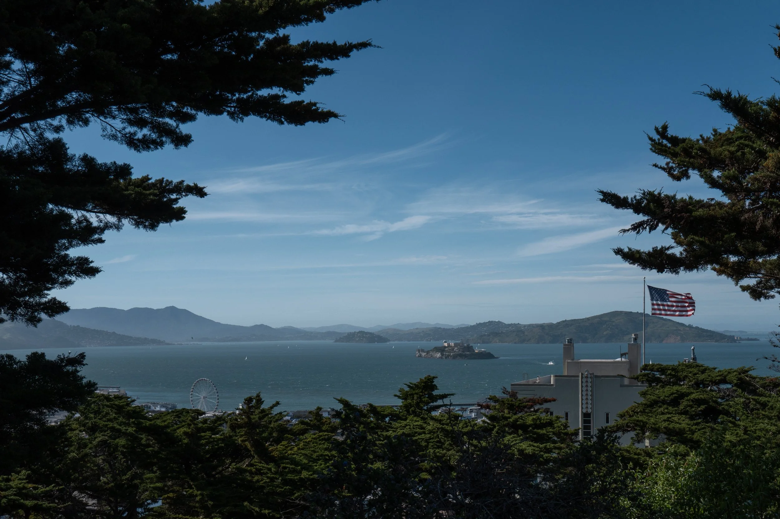 Vue d'une baie avec la mer, une petite île avec un bâtiment, et un drapeau américain dans une zone arborée avec des montagnes en arrière-plan.