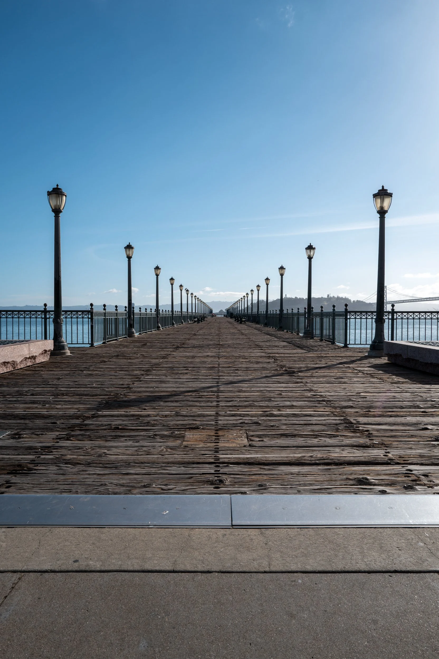 Un pont en bois avec des lampadaires alignés, surplombant une étendue d'eau, avec un ciel bleu clair.