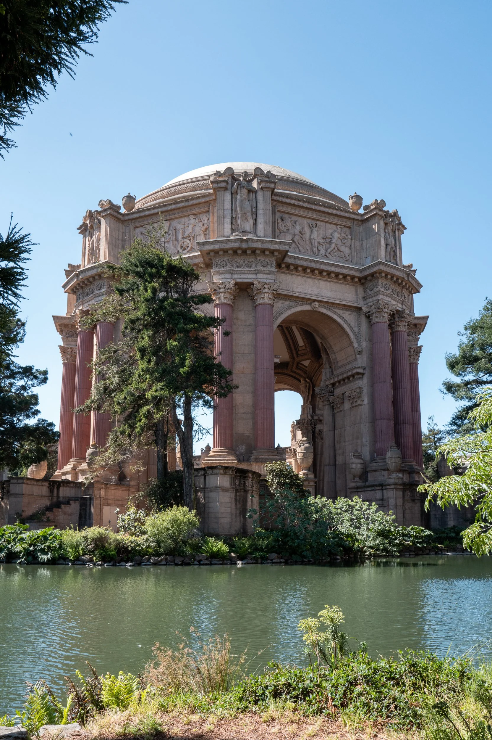 Une structure architecturale ronde et ancienne, avec des colonnes de couleur rose et des sculptures ornant le haut, entourée d'eau et de végétation