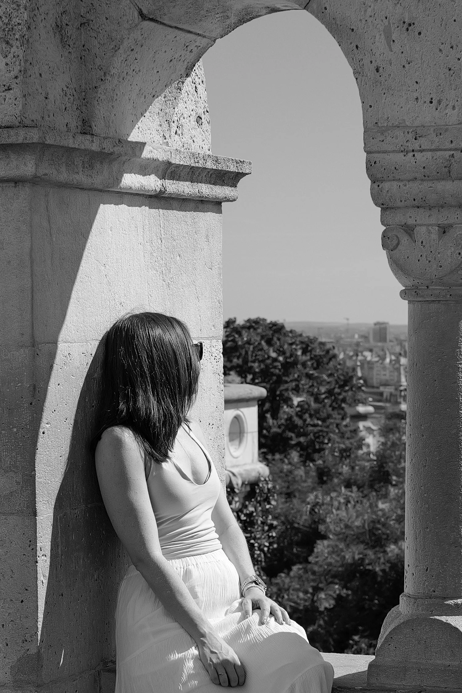 Une femme aux cheveux bruns longs et bouclés, portant un débardeur et une jupe longue, est assise sur un banc en pierre au soleil, dans un lieu historique avec des colonnes. Il y a des arbres et une ville en arrière-plan.