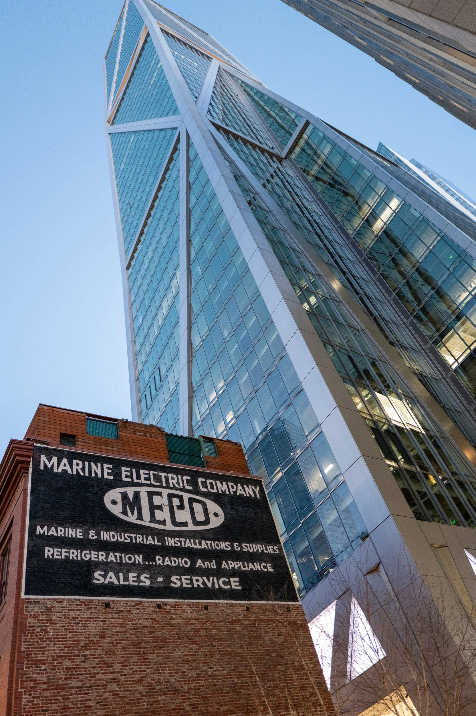 Un bâtiment moderne en verre et en acier vu d'en bas, avec une vieille façade en briques en dessous portant une publicité pour Marine Electric Company.