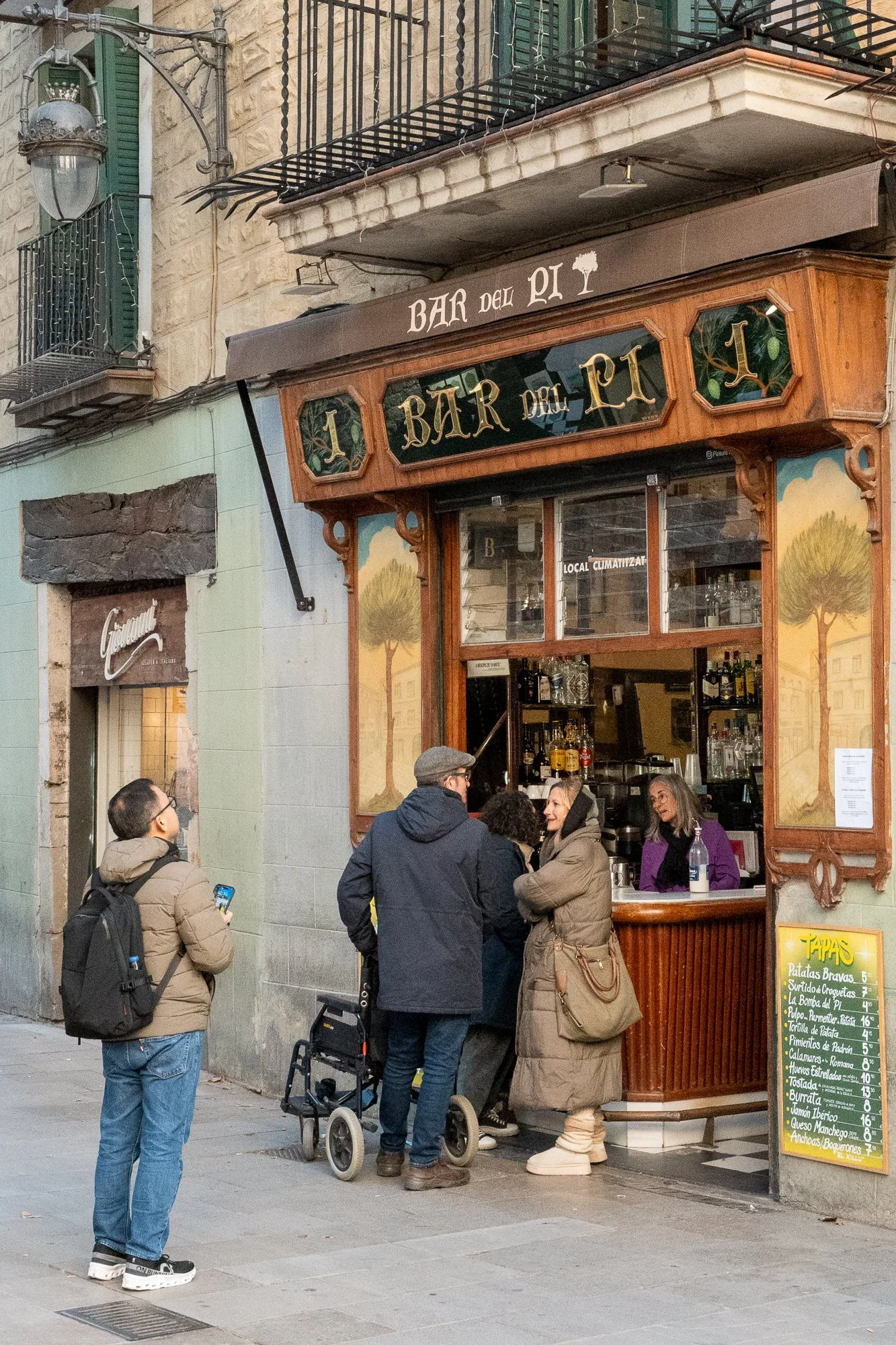 Un groupe de personnes fait la queue devant un bar appelé 'Bar del Pi' dans une rue de ville, avec un homme prenant une photo ou vidéo avec son téléphone.