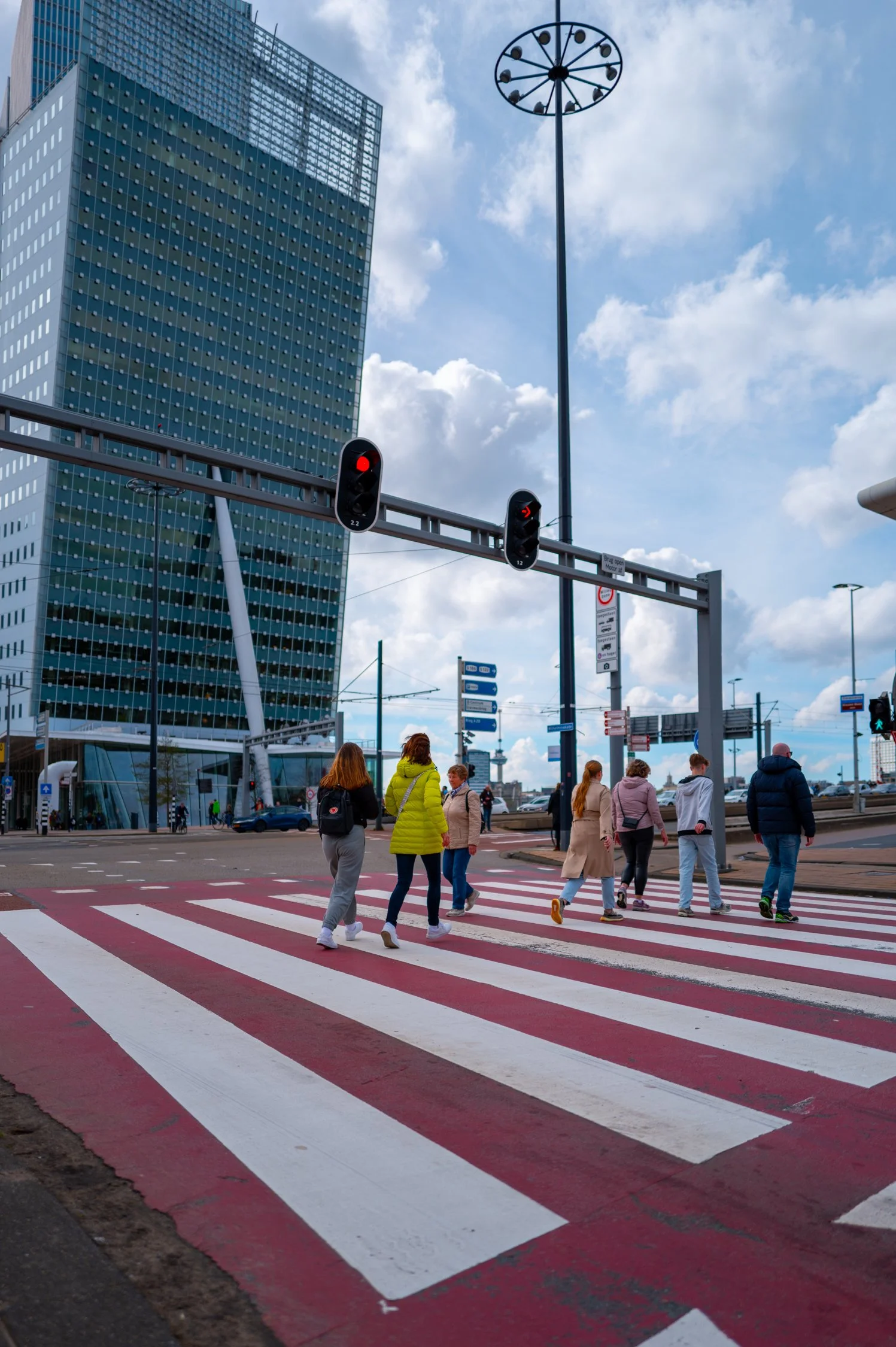 Groupe de personnes traversant un passage piéton dans une ville moderne, avec un bâtiment en verre et un ciel partiellement nuageux en arrière-plan.