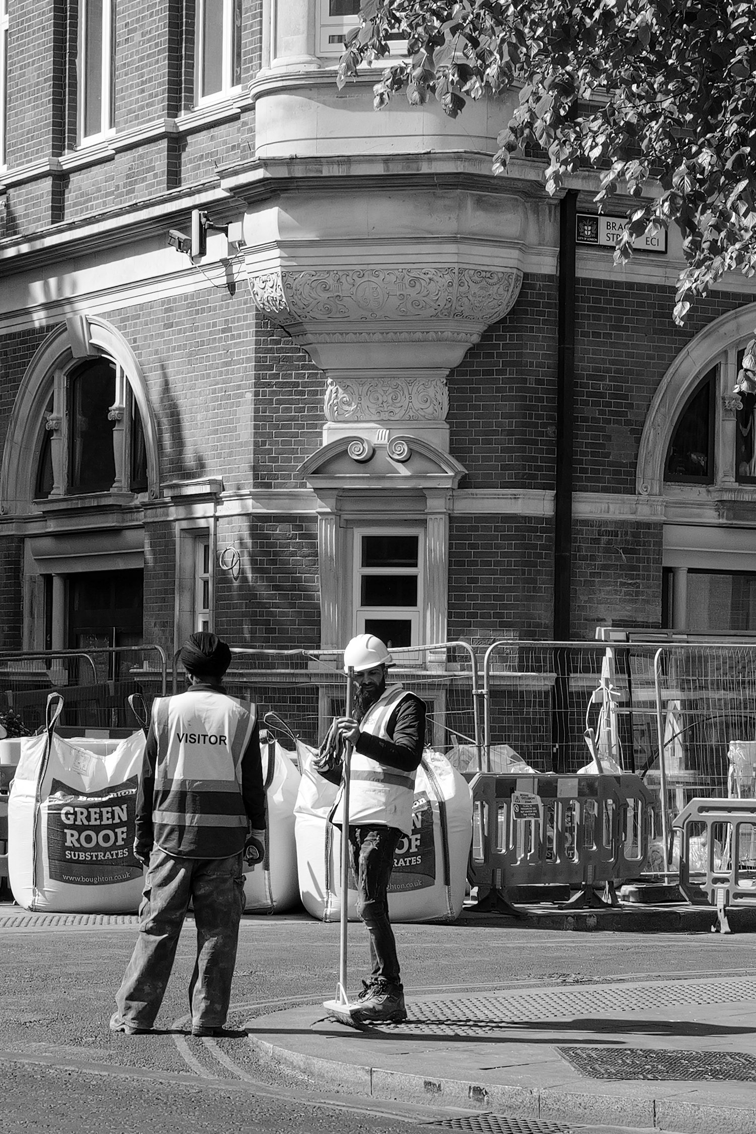 Vue de deux ouvriers de construction avec des gilets de sécurité, au travail devant une barrière de sécurité, avec un bâtiment en briques en arrière-plan.