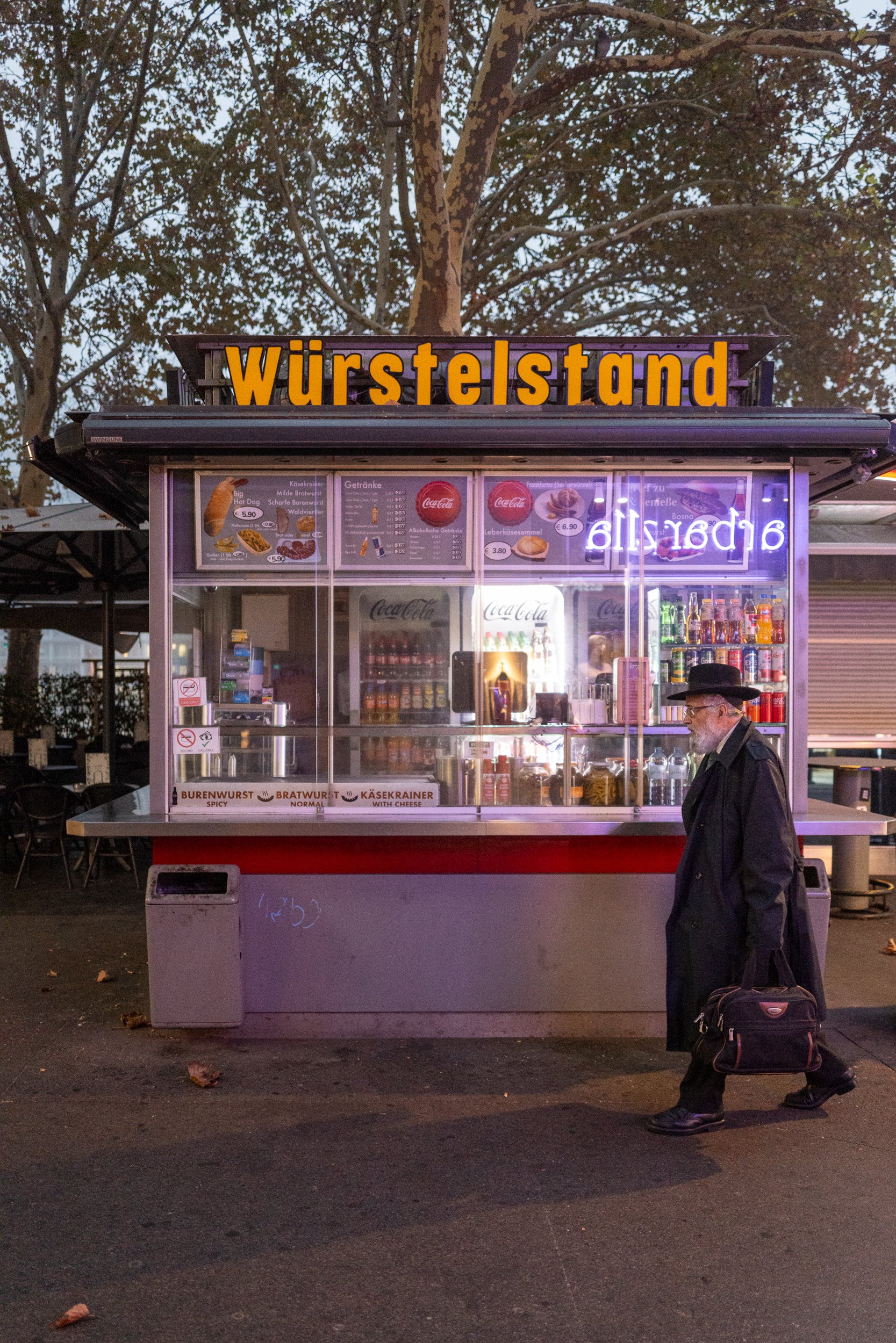 Un vendeur de hot dogs devant un stand appelé 'Würstelstand' avec des affiches de menus et des boissons. Une personne avec un manteau noir, un chapeau noir, des lunettes, portant un sac, passe devant le stand en soirée.