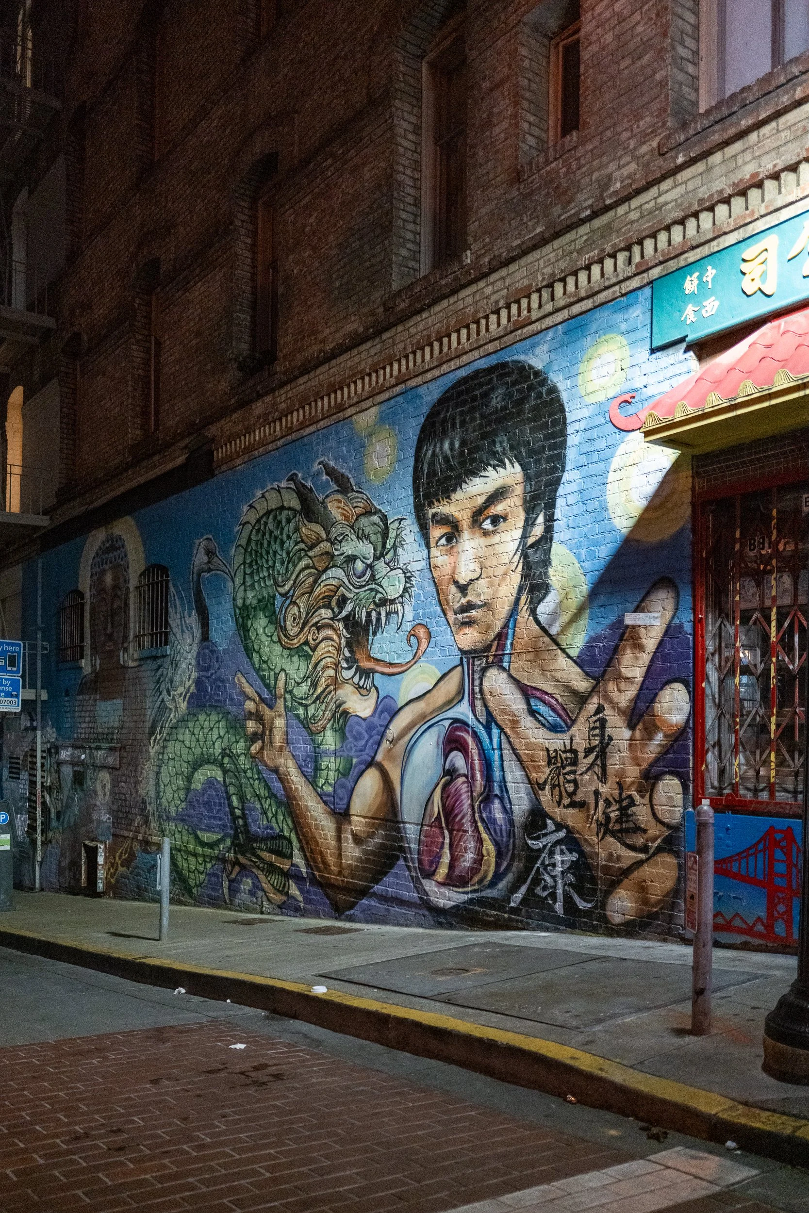 Graffiti mural représentant un jeune homme avec une main en forme de dragon et des caractères chinois sur un mur en briques dans une rue urbaine la nuit.