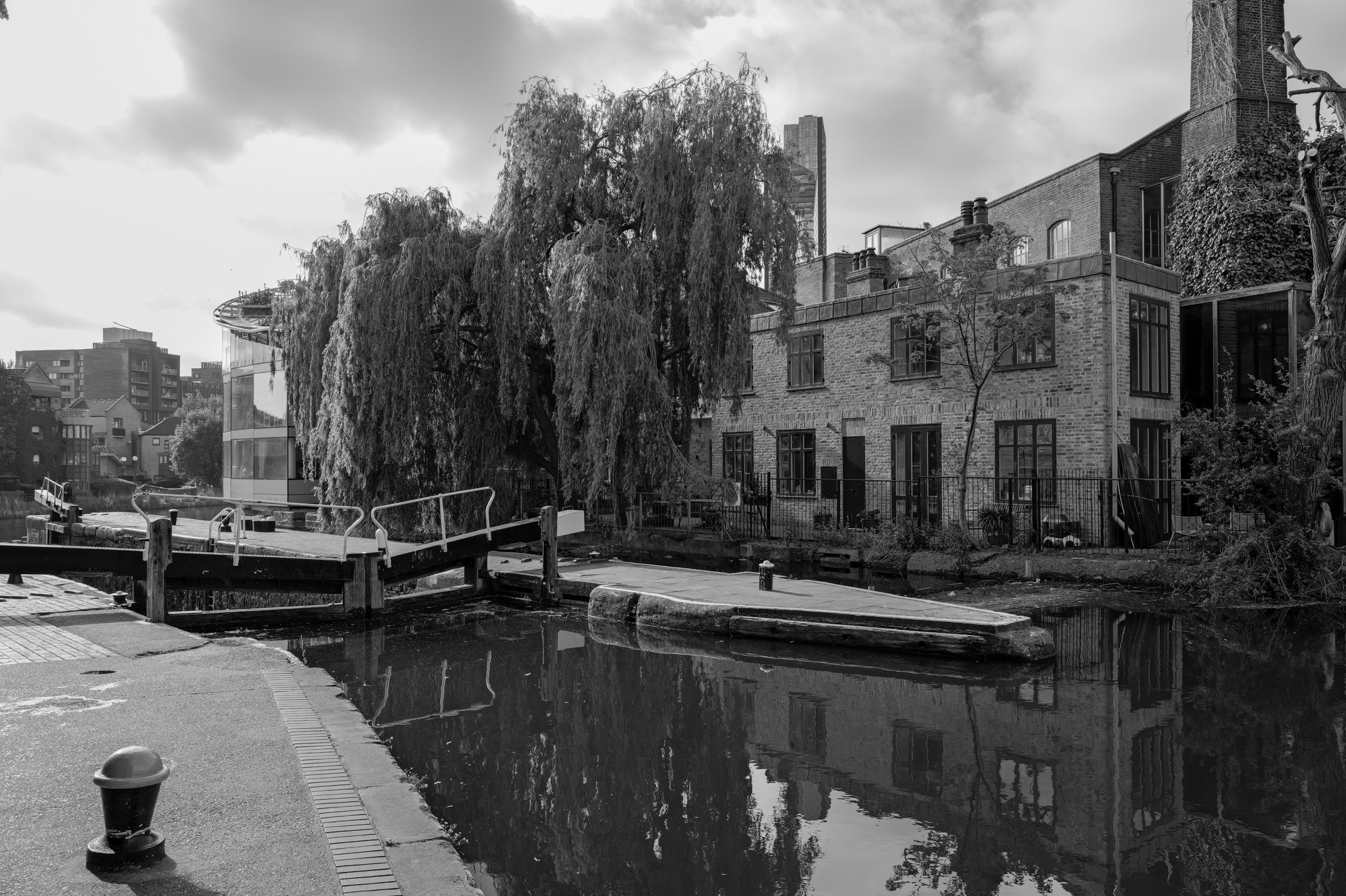 Vue d'une scène urbaine en noir et blanc avec un bâtiment en briques, une grande arbre, un canal ou étang, et un pont en bois. Des bâtiments modernes et un ciel nuageux en arrière-plan.