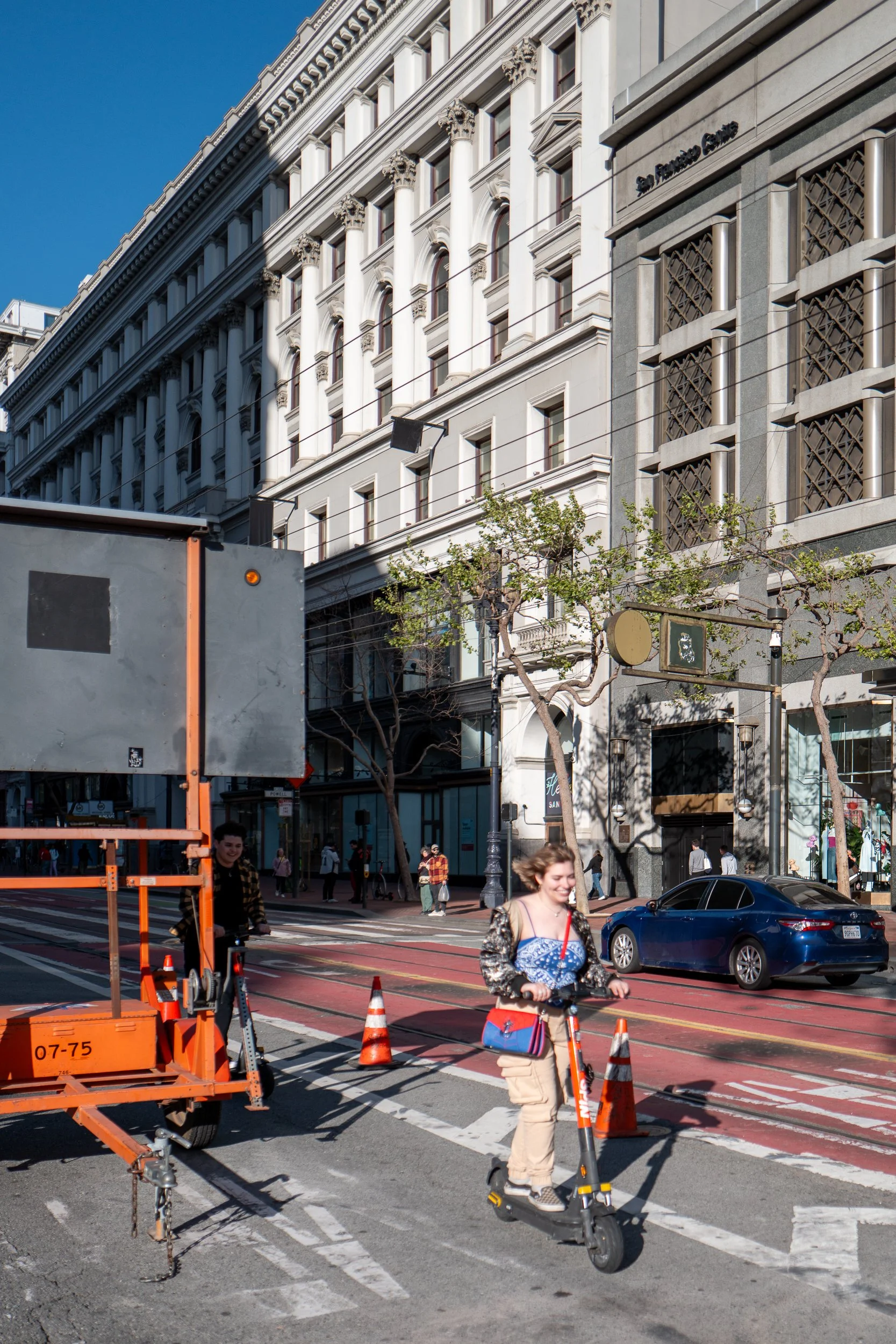 Une jeune femme souriante circule à scooter à un passage piéton dans une rue urbaine. En arrière-plan, un bâtiment ancien et un véhicule noir sont visibles.