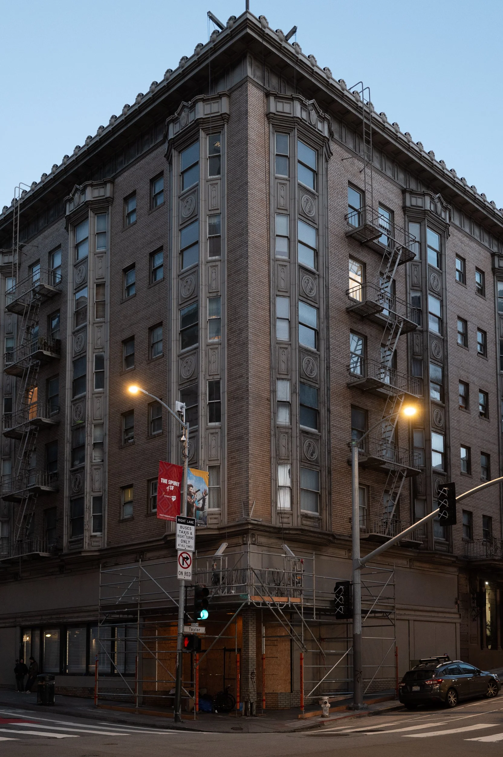 Immeuble en briques avec fenêtres et escaliers de secours extérieurs, situé à un coin de rue, avec des lampadaires allumés en soirée.
