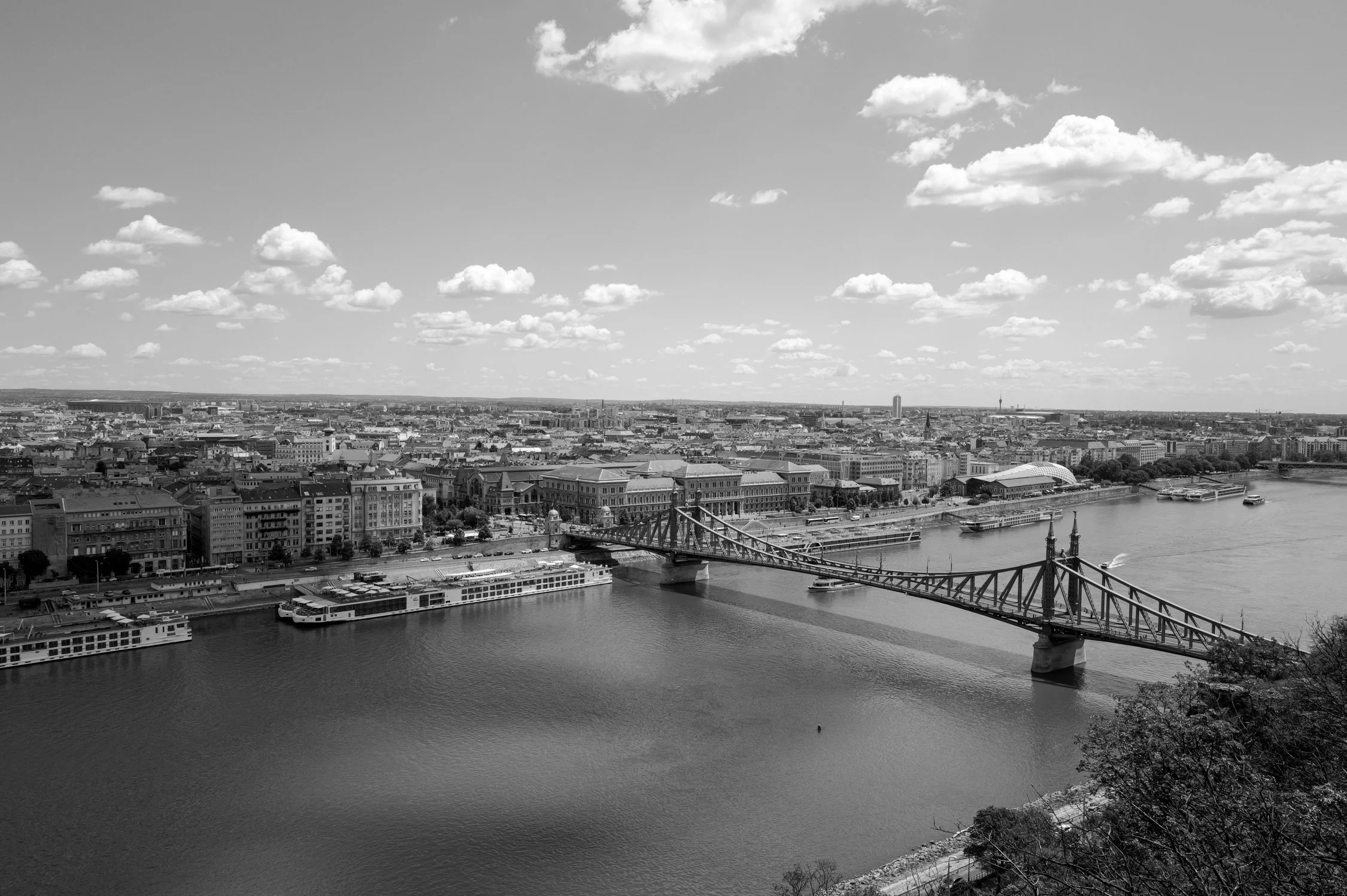 Vue aérienne d'une ville avec un fleuve et un pont suspendu, avec des bâtiments urbains, des nuages dans le ciel et des bateaux sur l'eau.