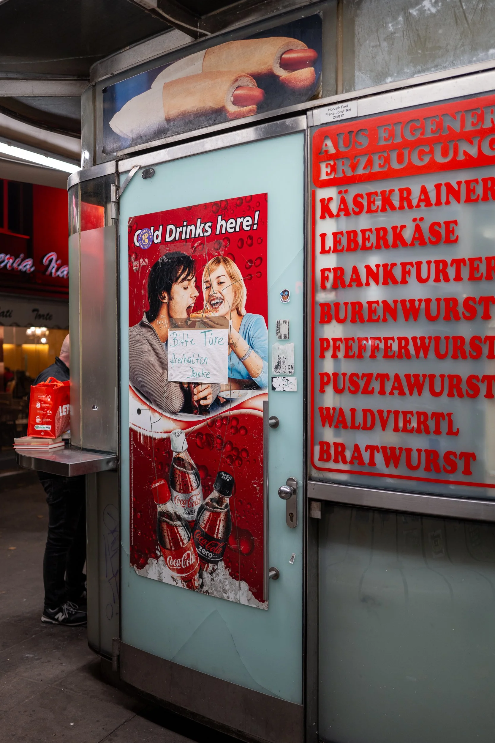 Stall de nourriture avec des affiches en allemand et en anglais, montrant des boissons Coca-Cola et des petits plats, avec deux personnes partageant un moment joyeux.