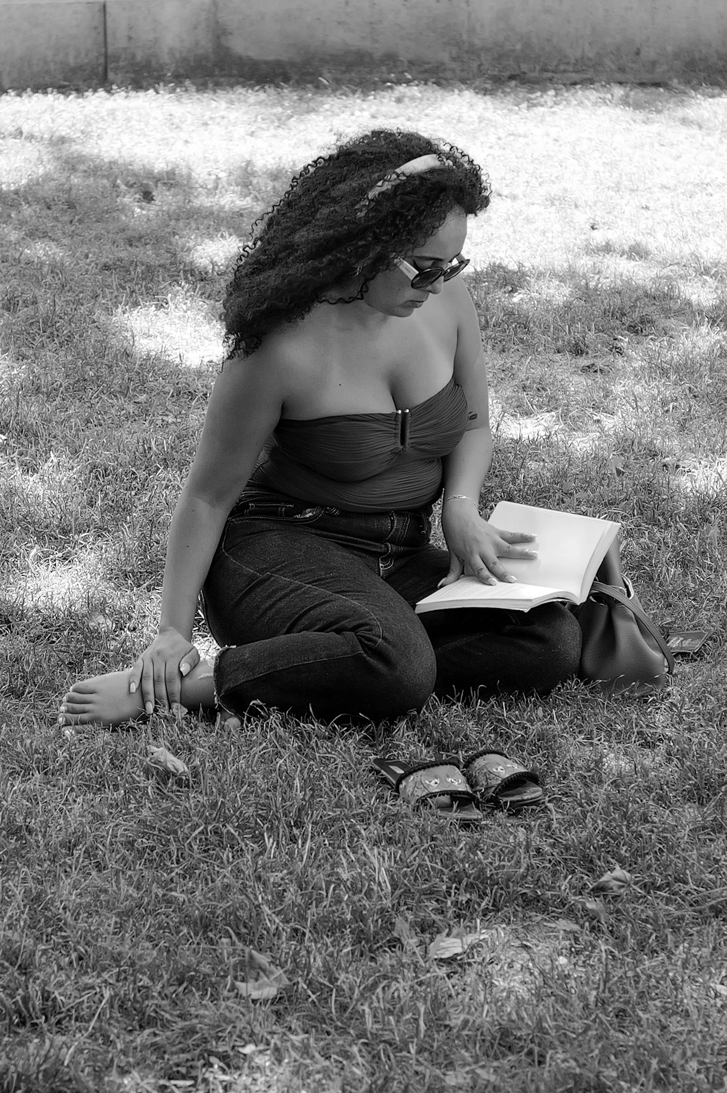 Une femme assise sur l'herbe, portant un haut sans manches avec une fermeture à l'avant, des lunettes de soleil, lisant un livre, avec des chaussures à fleurs posées devant elle.