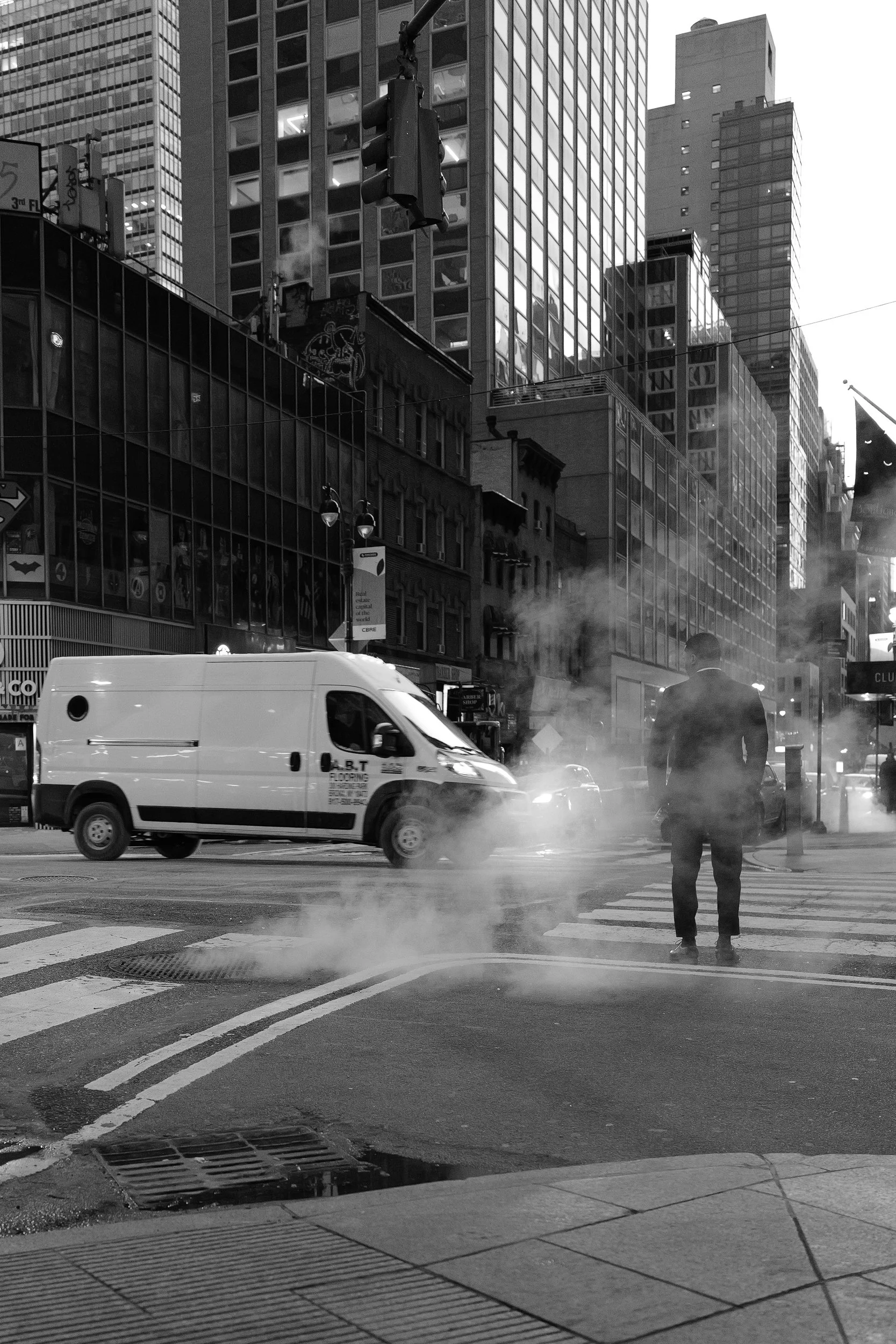 Une rue urbaine animée avec des gratte-ciel, un véhicule blanc et une personne en costume traversant un passage piéton, de la vapeur ou de la fumée s'échappant du sol en mode noir et blanc.