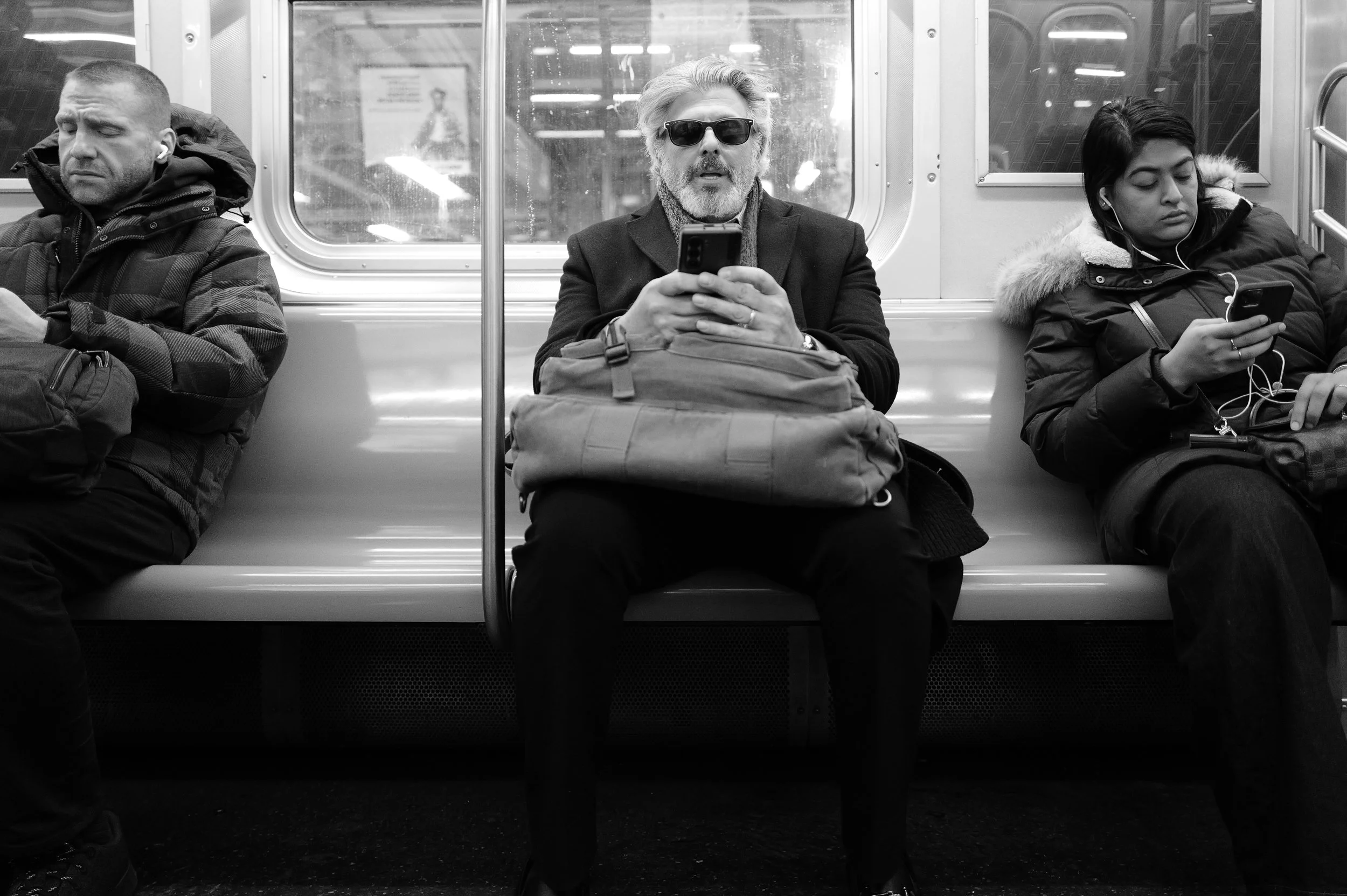 Trois personnes assises dans le métro, toutes utilisant leur téléphone portable. L'homme au centre porte des lunettes de soleil et un manteau foncé, alors que les deux autres portent des vêtements chauds.
