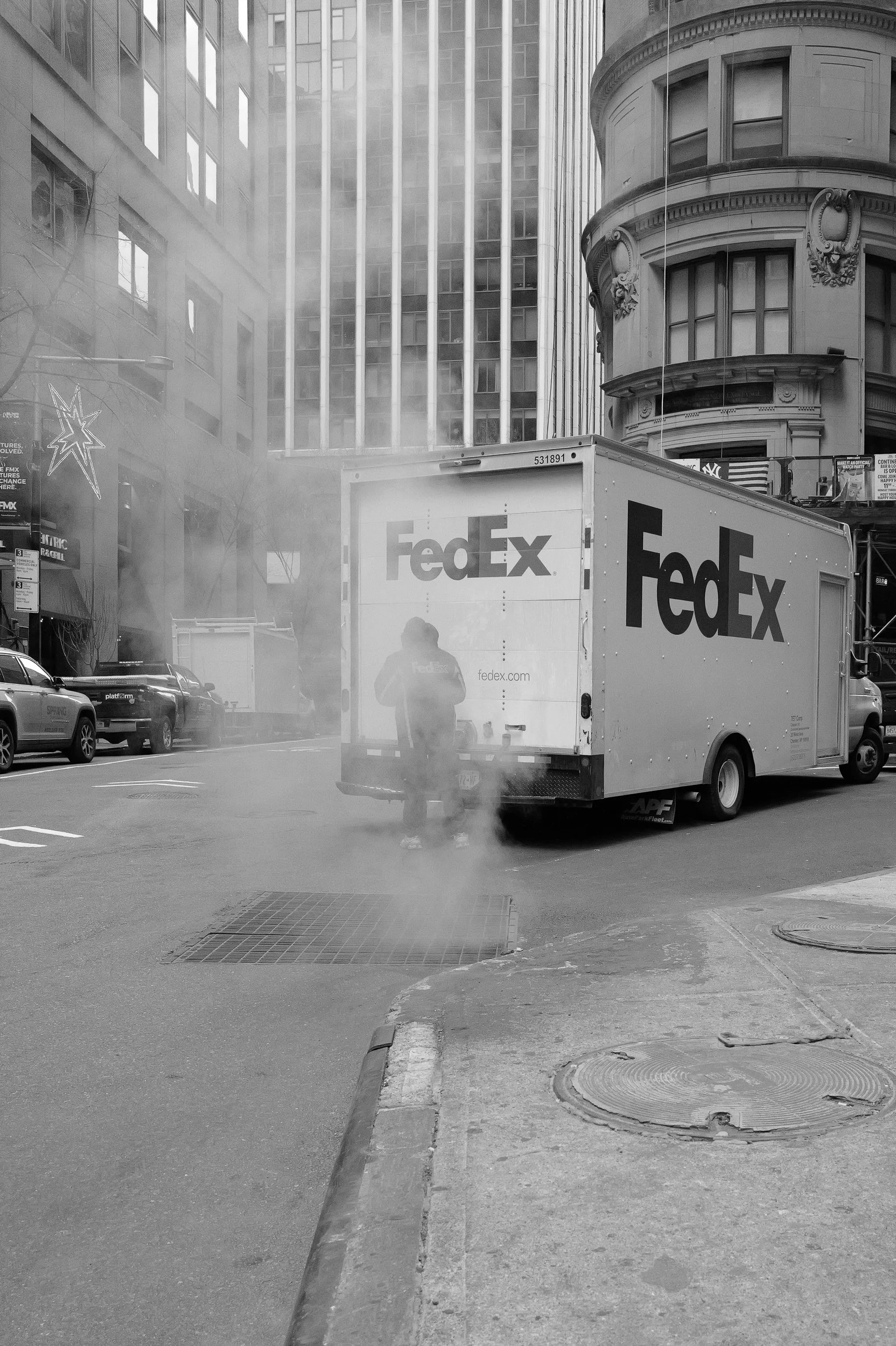 Camion de livraison FedEx dans une rue urbaine, avec de la vapeur ou de la fumée qui s'élève, en noir et blanc.