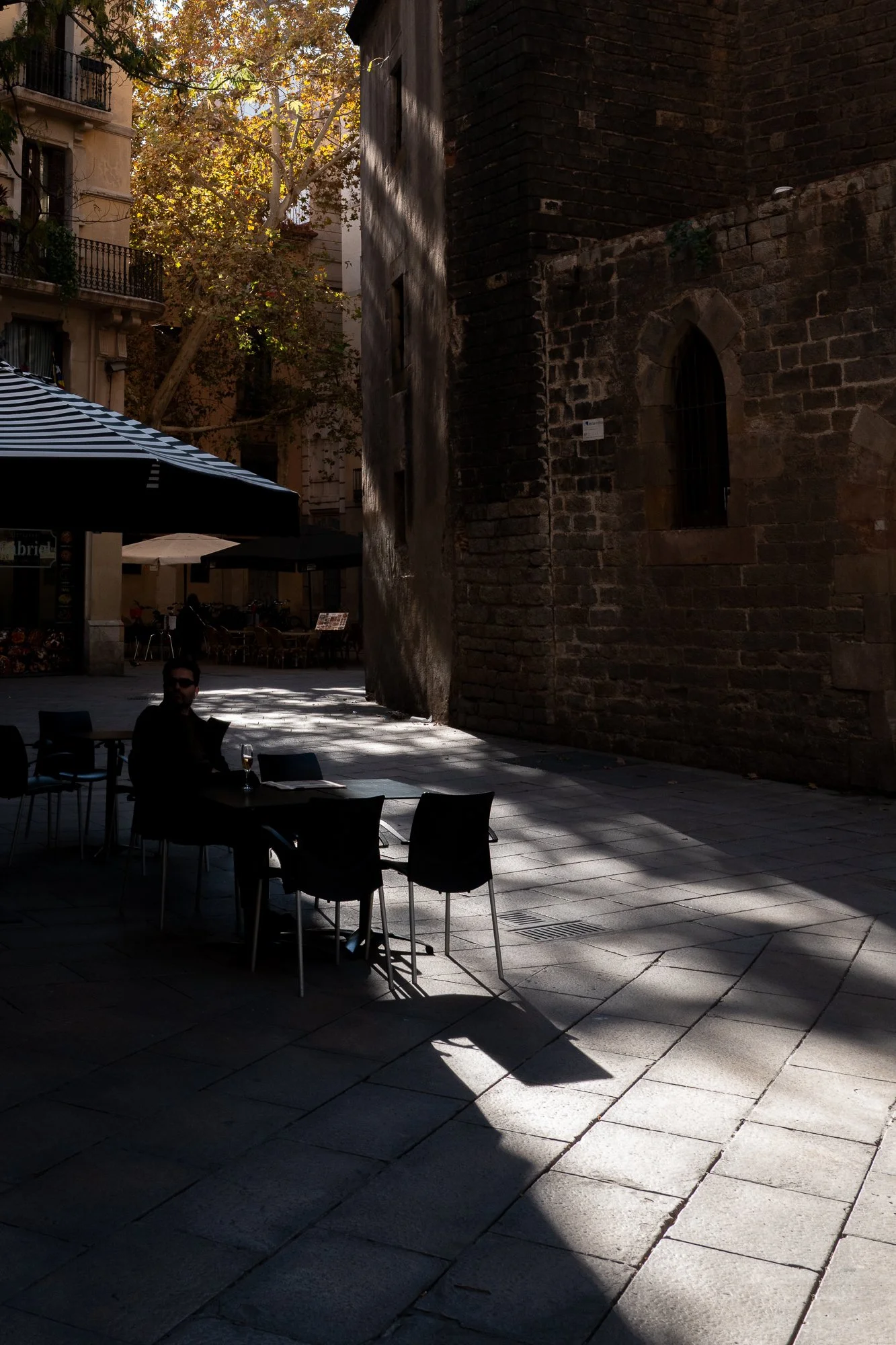 Une scène urbaine avec une terrasse de café en plein air, ombragée par des parasols rayés et entourée d'arbres aux feuilles jaunes, avec un homme assis à une table portant des lunettes de soleil. Le bâtiment ancien en pierre est en arrière-plan, avec