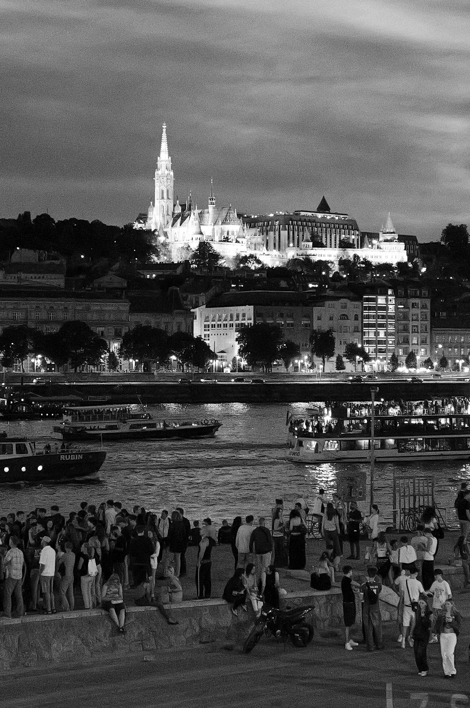 Une scène nocturne avec une foule de personnes au bord de l'eau, des bateaux naviguant sur la rivière, et un bâtiment historique illuminé au sommet d'une colline en arrière-plan.