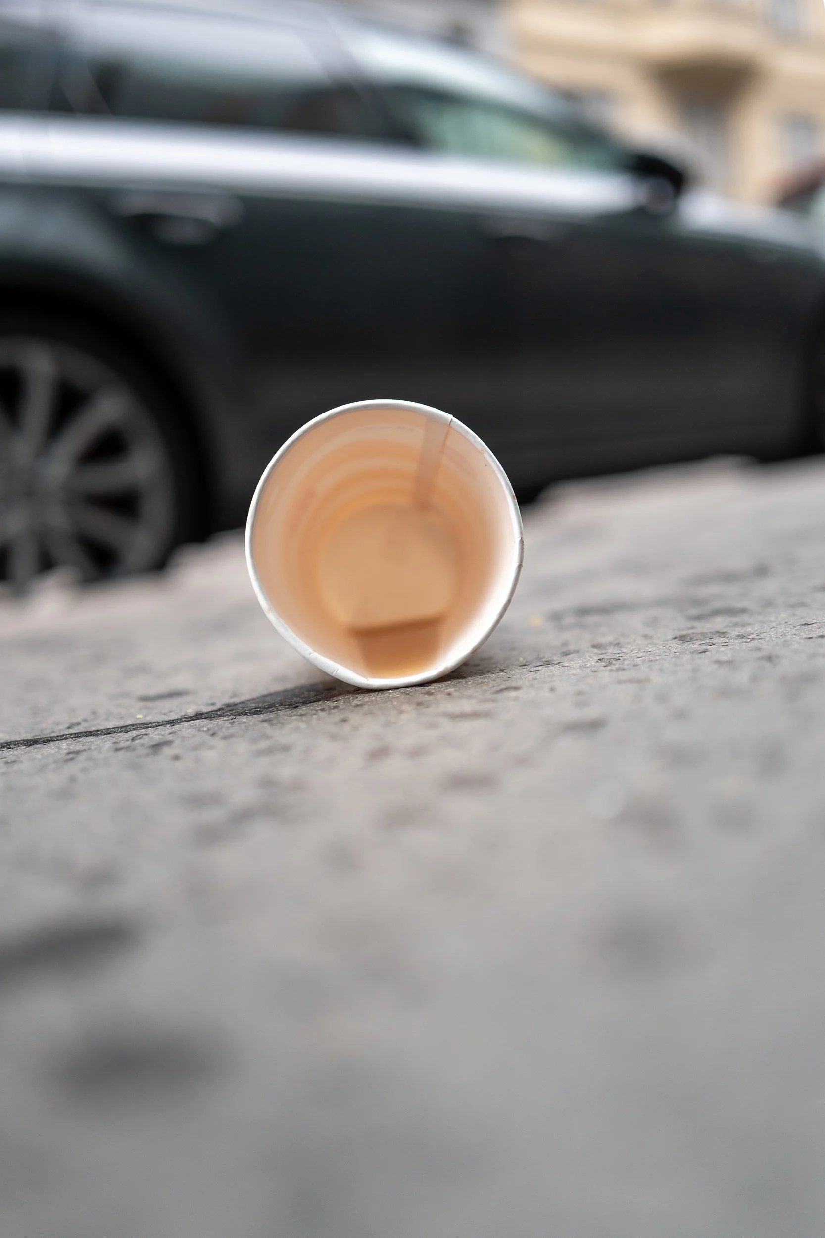 Un gobelet en papier vide renversé sur un trottoir en béton avec une voiture noire floue en arrière-plan.