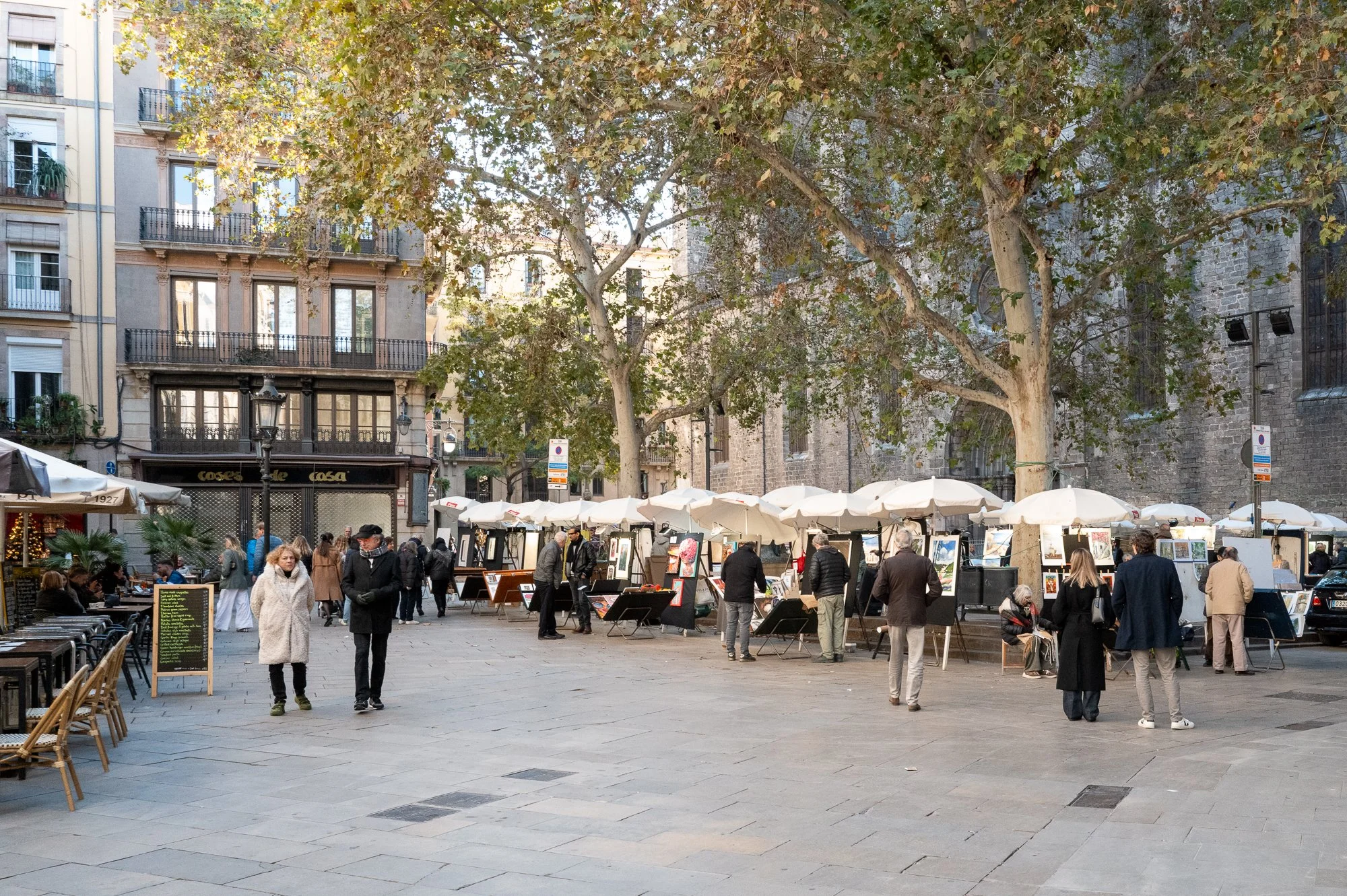 Marché d'art en plein air avec stands sous des parasols blancs, personnes qui regardent des œuvres, place pavée, grands arbres, bâtiments anciens en arrière-plan.