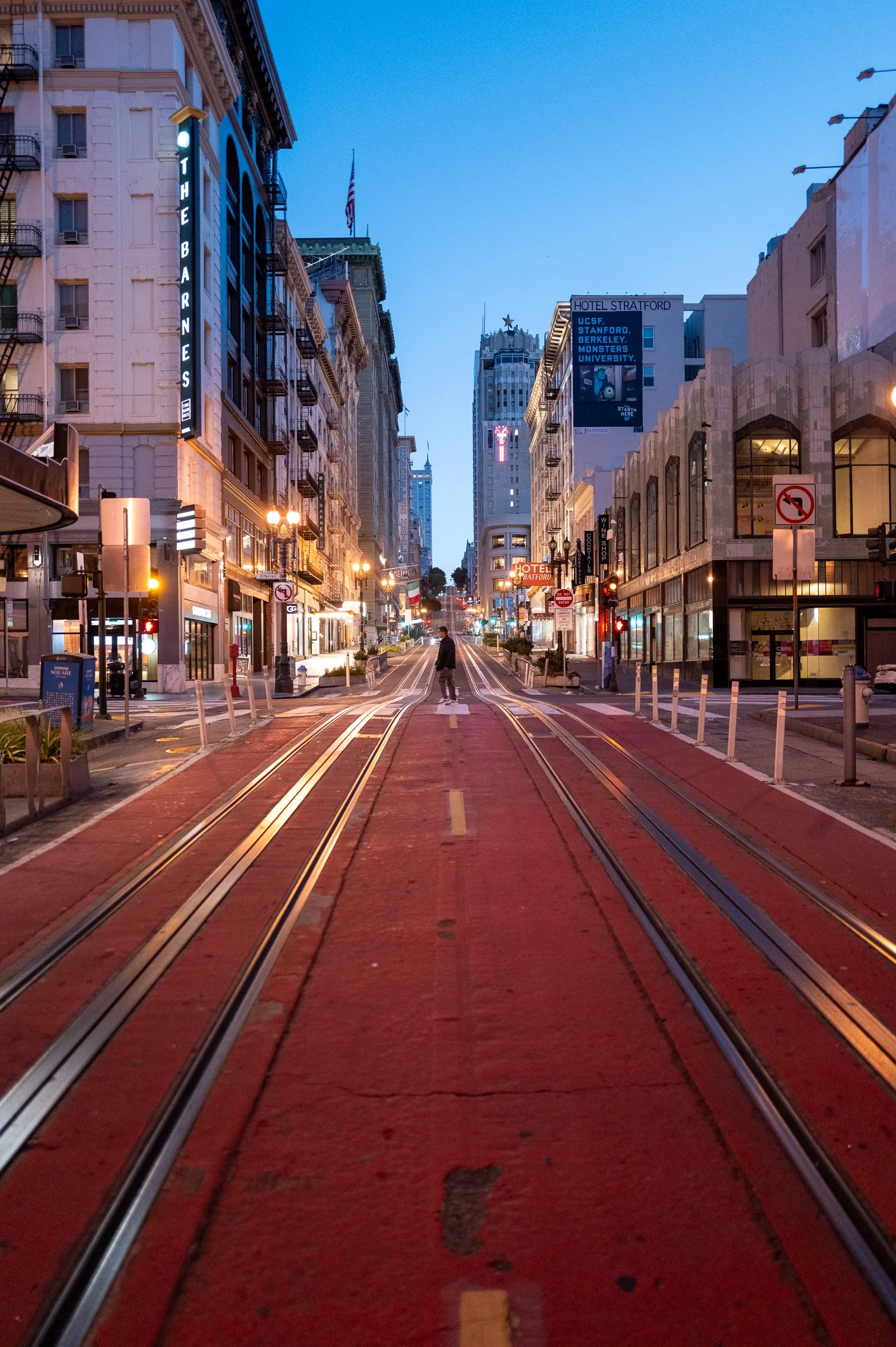 Une rue urbaine avec des rails de tramway rouges au centre, des bâtiments hauts et éclairés de chaque côté, et un ciel crépusculaire. Il y a un piéton au milieu de la voie. Des panneaux publicitaires et de signalisation sont visibles.