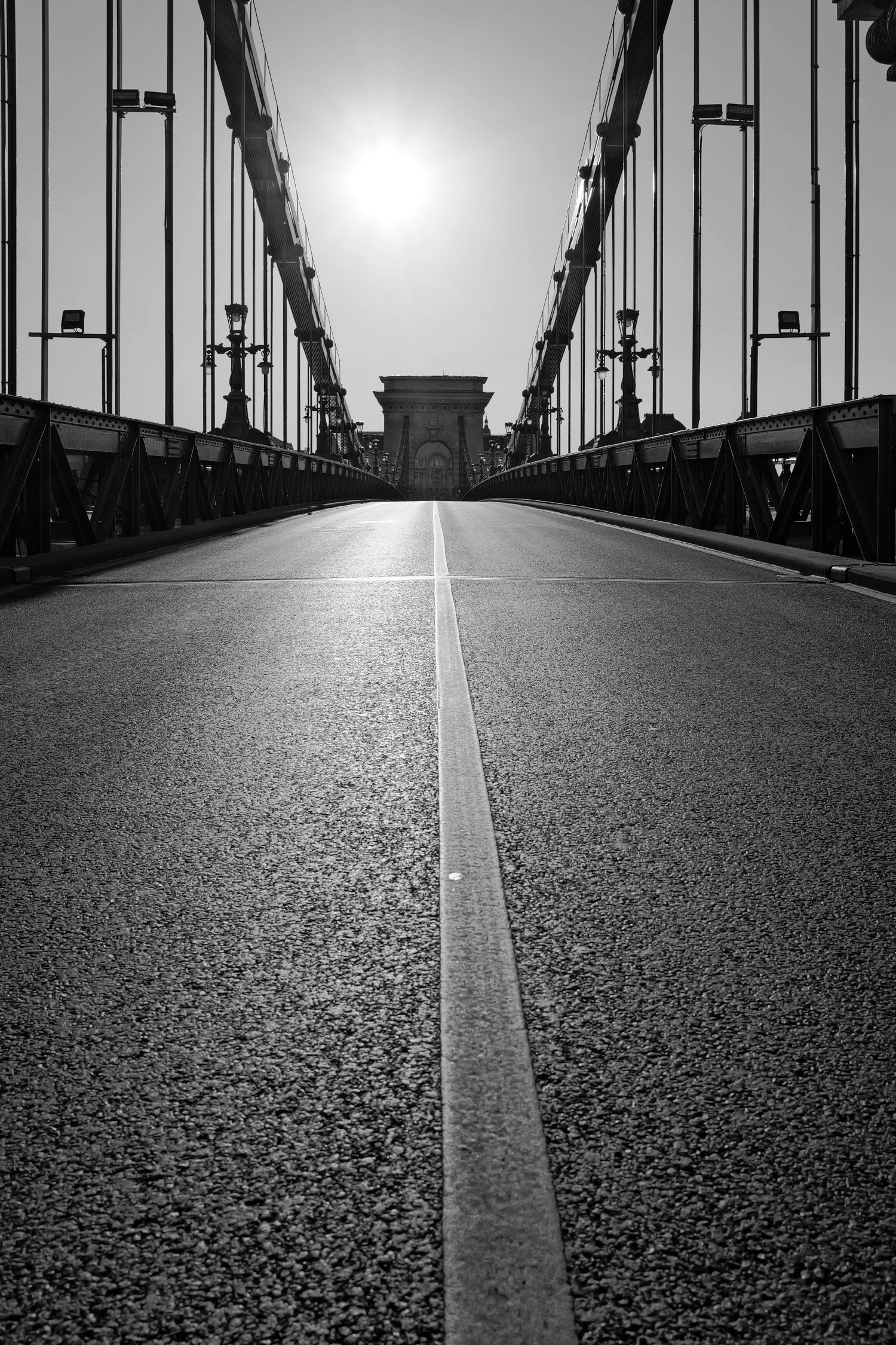 Photo en noir et blanc d'un pont suspendu avec une voie au centre, des lampadaires sur les côtés, et un bâtiment au fond sous un soleil brillant dans un ciel clair.