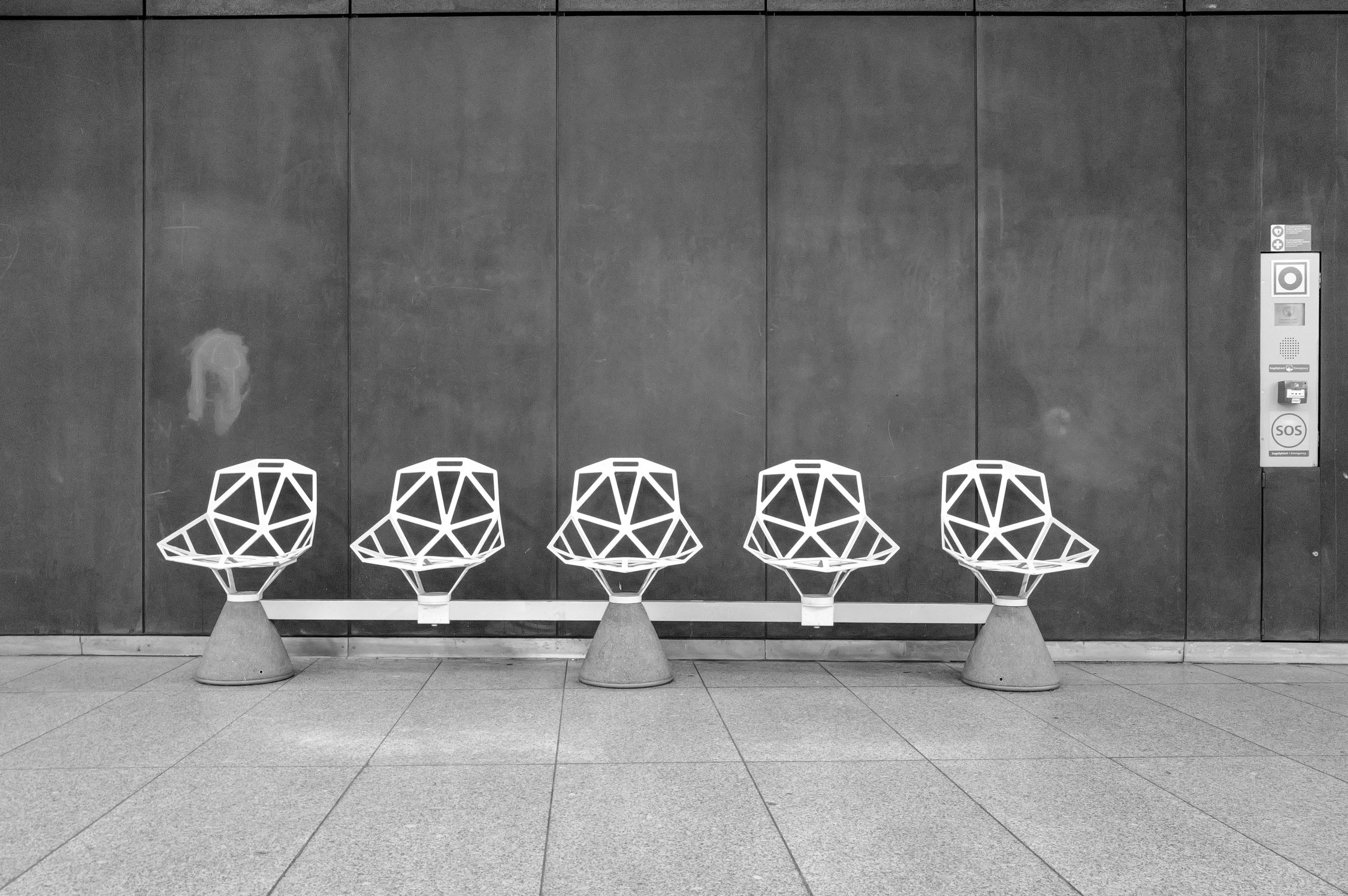 Quatre fauteuils en forme de géométrie blanche, alignés contre un mur en pierre grise, dans une salle d'attente ou un espace public.