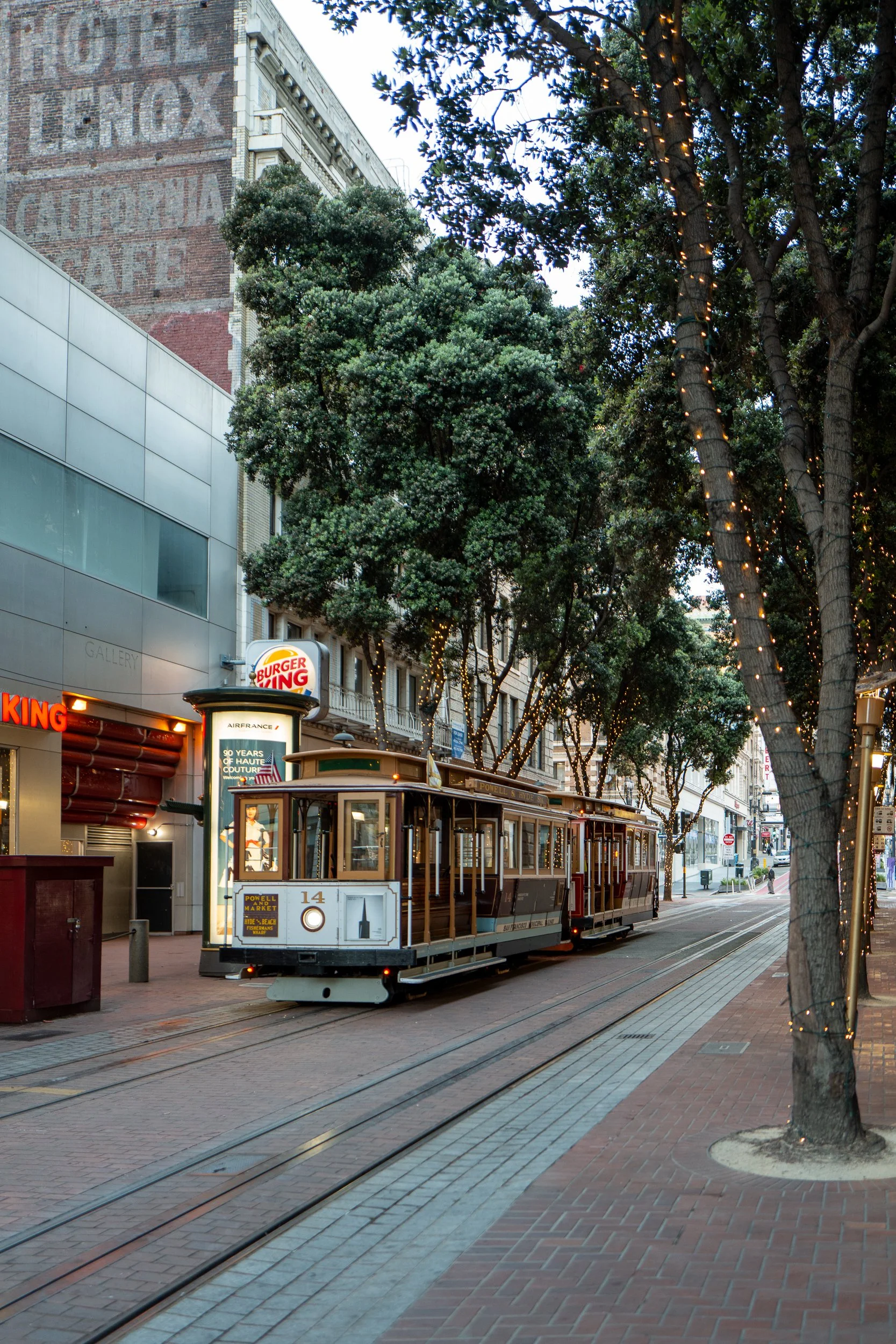 Un tramway vintage circule dans une rue urbaine bordée d'arbres et de bâtiments, avec un panneau Burger King visible en arrière-plan.