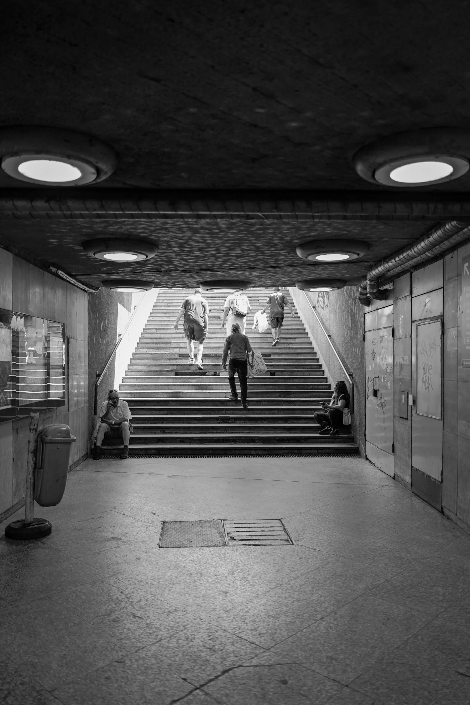 Une scène en noir et blanc montrant un groupe de personnes montant des escaliers à l'extérieur, sous un passage couvert. Deux femmes sont assises sur le sol à gauche et à droite, une parlant au téléphone et l'autre utilisant un téléphone portable.