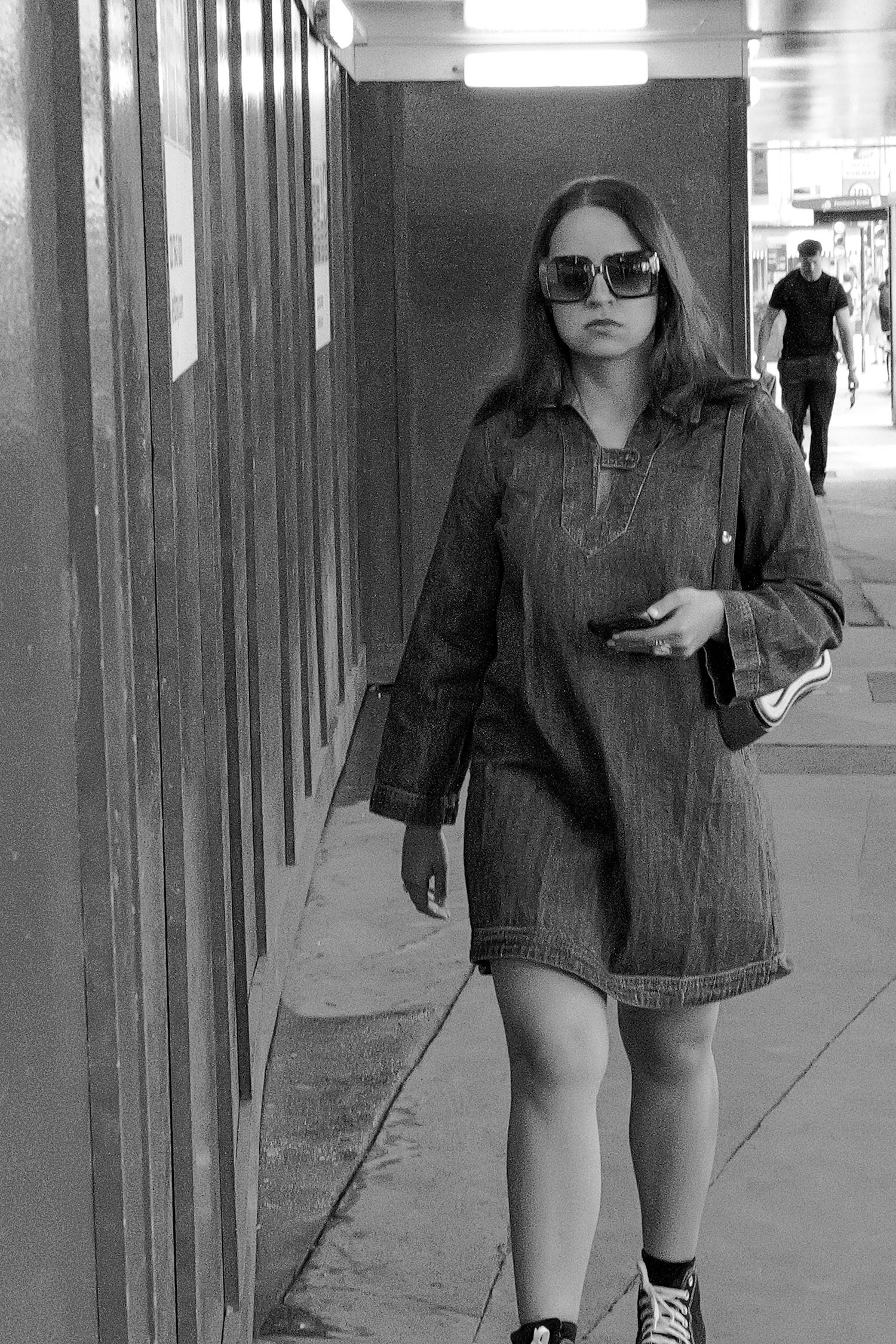 Femme marchant dans la rue portant des lunettes de soleil, une veste en jean, une robe courte, et des baskets, en noir et blanc.