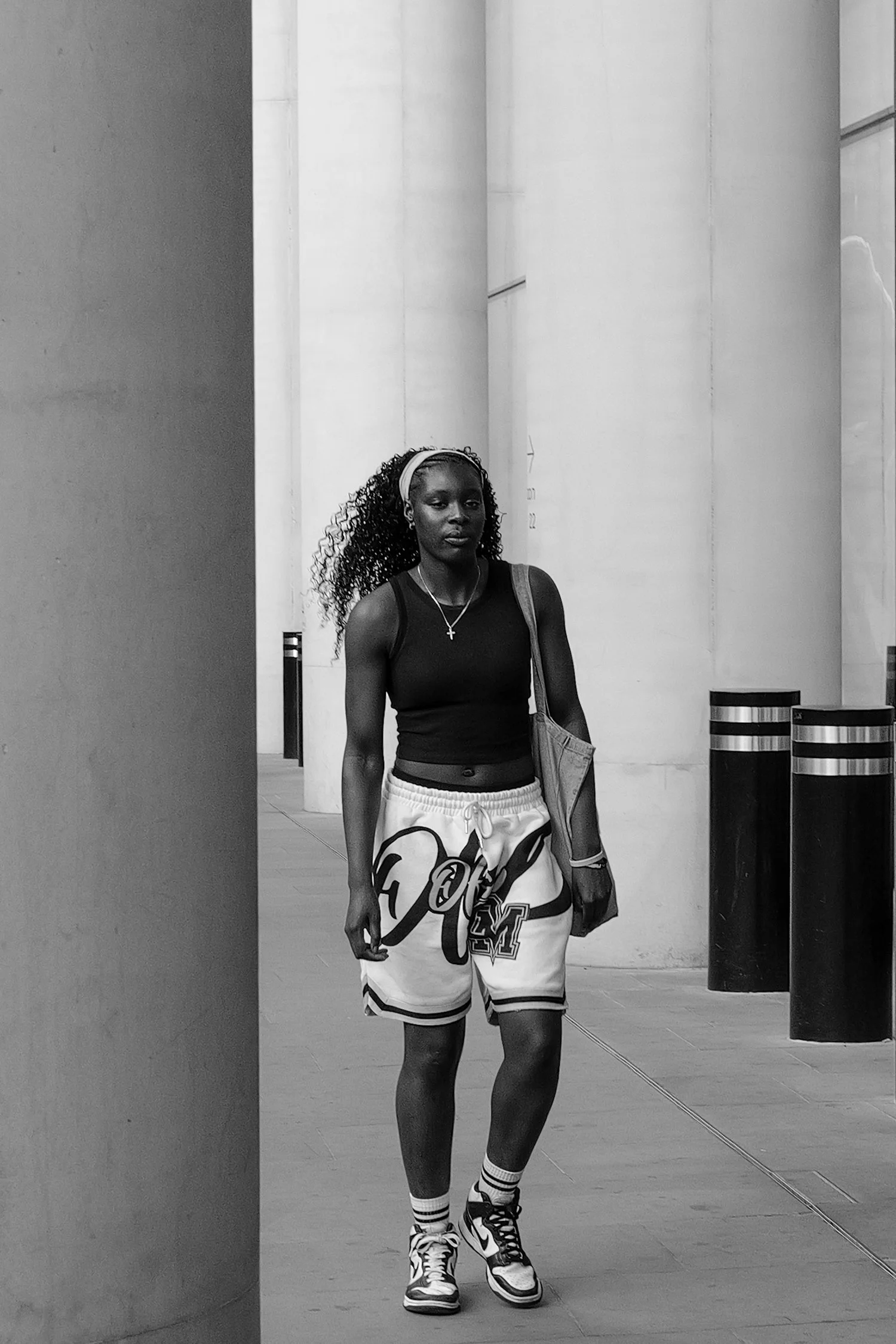 Une jeune femme portant un short de basketball, un haut noir sans manches, des chaussures de sport, des chaussettes à rayures et un sac à la main, marche dans un espace intérieur minimaliste avec des colonnes blanches.