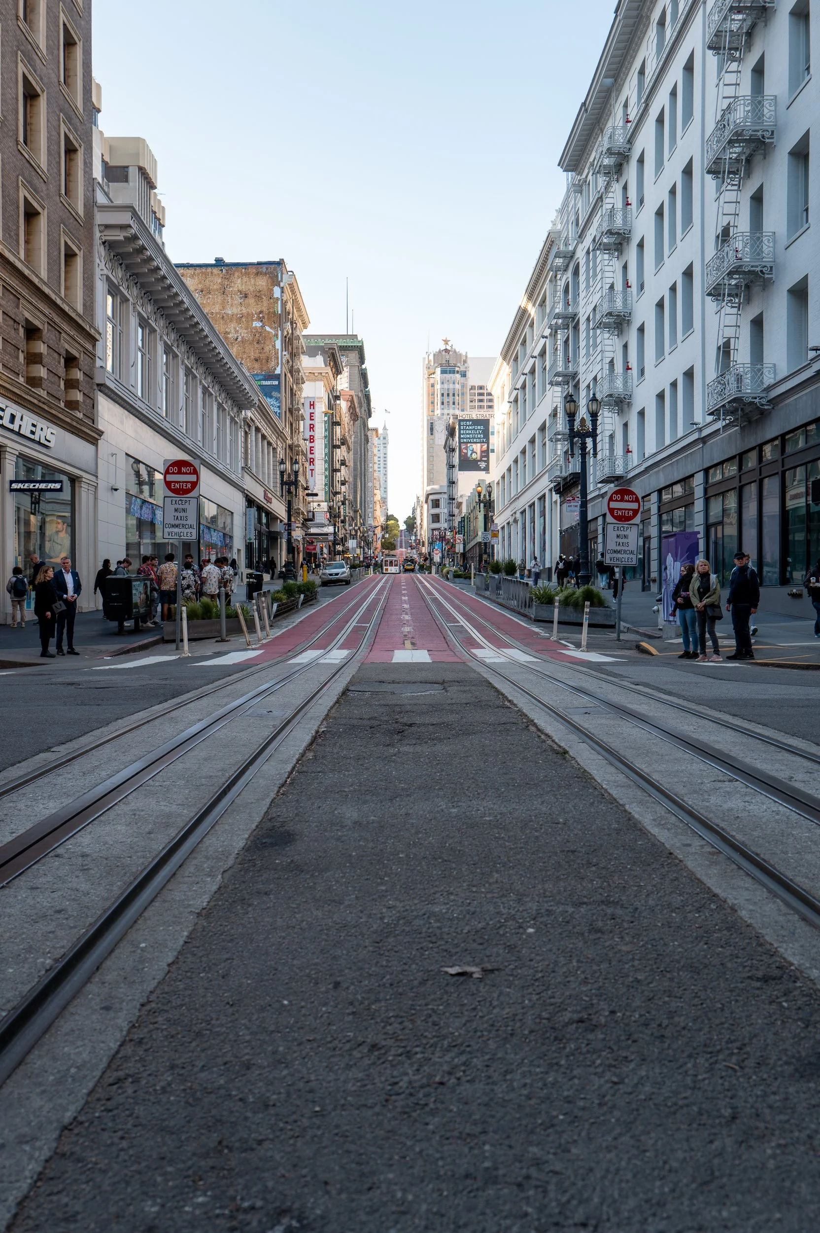 Une rue urbaine en ville avec des bâtiments de chaque côté et des rails de tramway au centre. Des piétons attendent au bord de la rue.