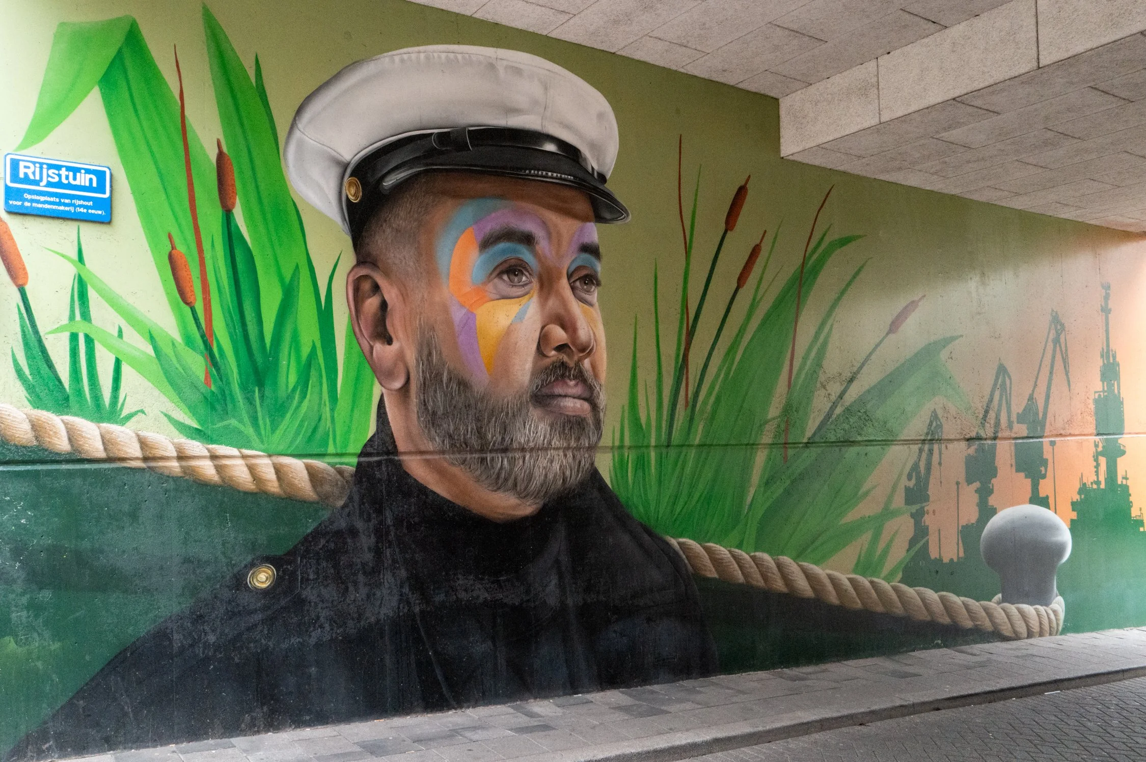 Mural coloré représentant une figure masculine portant un chapeau de marin, avec un maquillage coloré sur le visage, derrière des herbes vertes et des éléments architecturaux en ville.