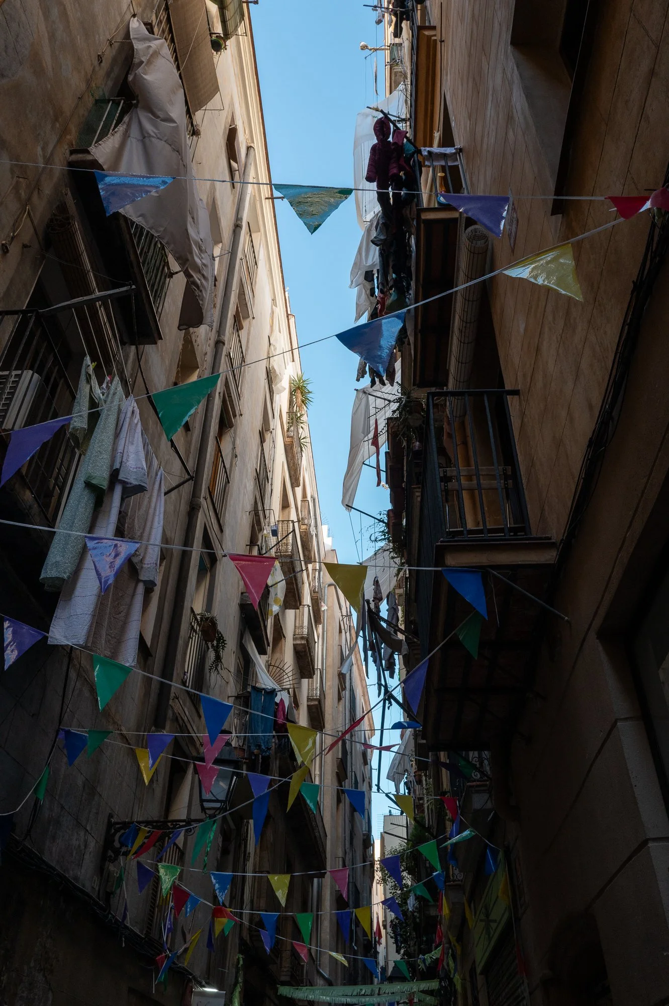 Ruelles étroites avec des bâtiments en pierre ou en béton, décorées avec des banderoles colorées en treillis, des vêtements suspendus pour sécher, et des balcons avec plantes et objets divers. Le ciel est visible au-dessus.