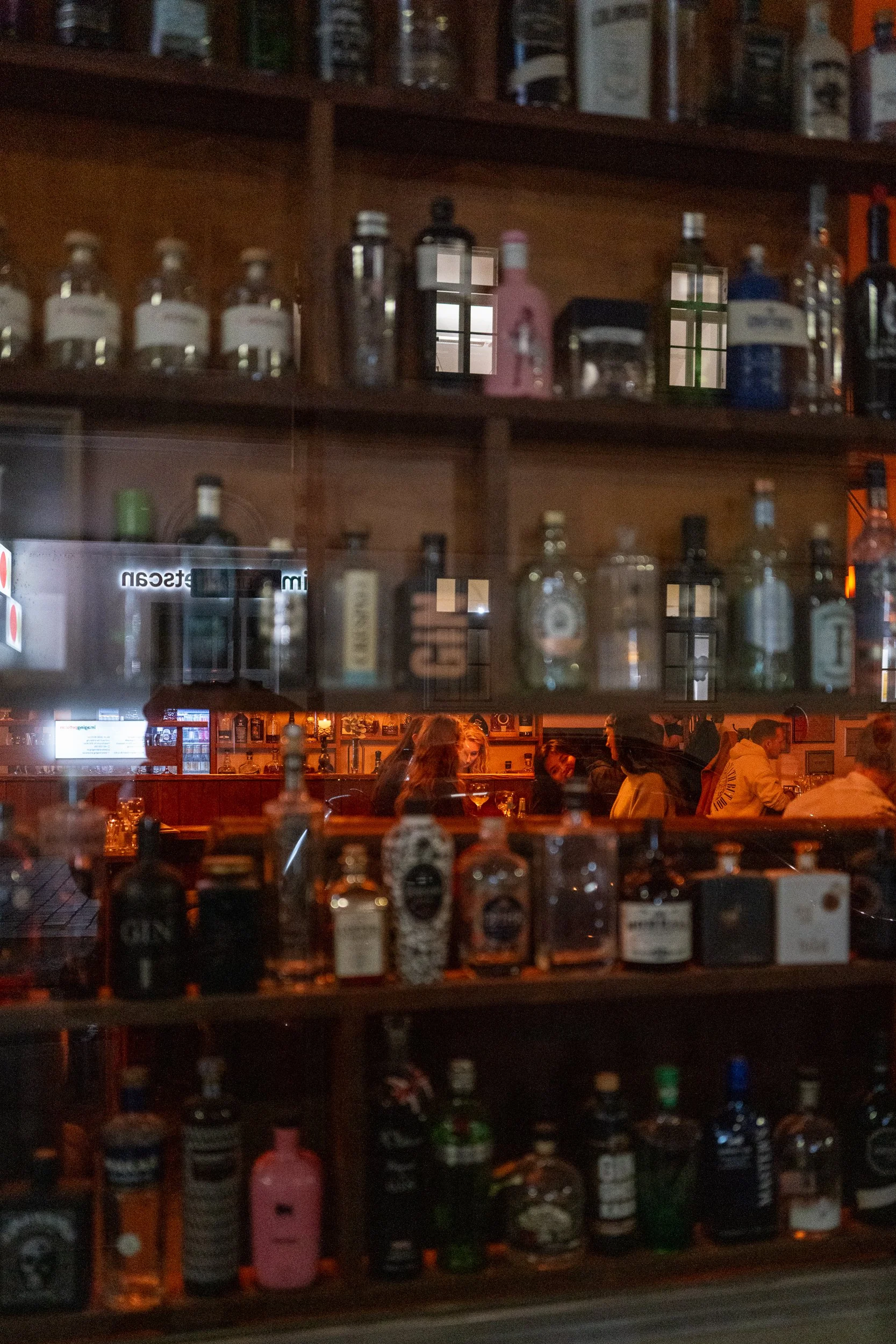 Une vue à travers une vitre d'un bar ou d'une salle de restaurant, avec des bouteilles d'alcool sur des étagères visibles à travers la vitre, et des personnes assises à une table au fond, dans une ambiance chaleureuse et animée.