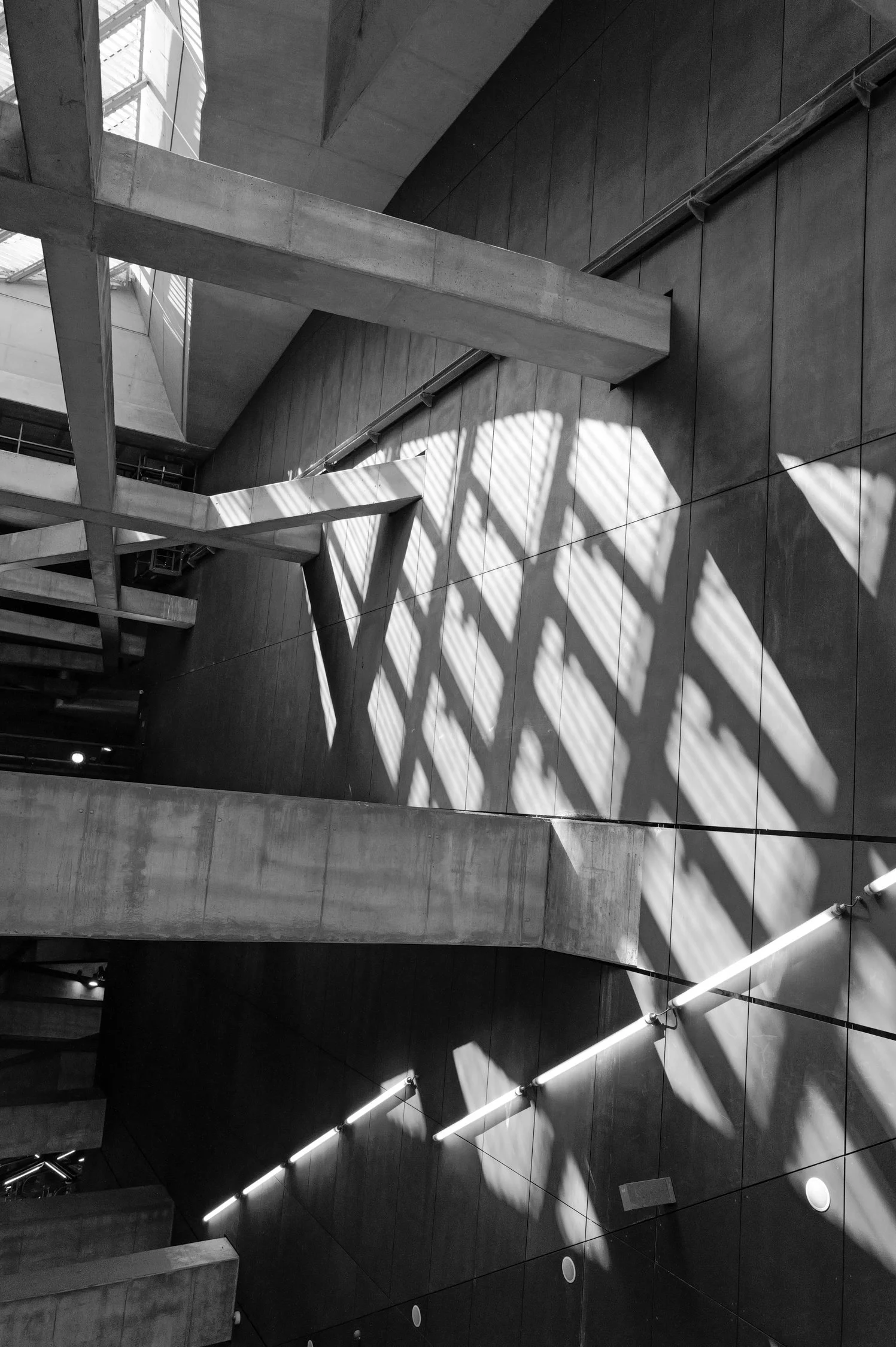 Une image en noir et blanc d'un intérieur moderne, présentant une structure architecturale avec des murs en béton, des escaliers, des piliers, des murs en panneaux, et des ombres provoquées par la lumière naturelle qui filtre à travers des fenêtres, 