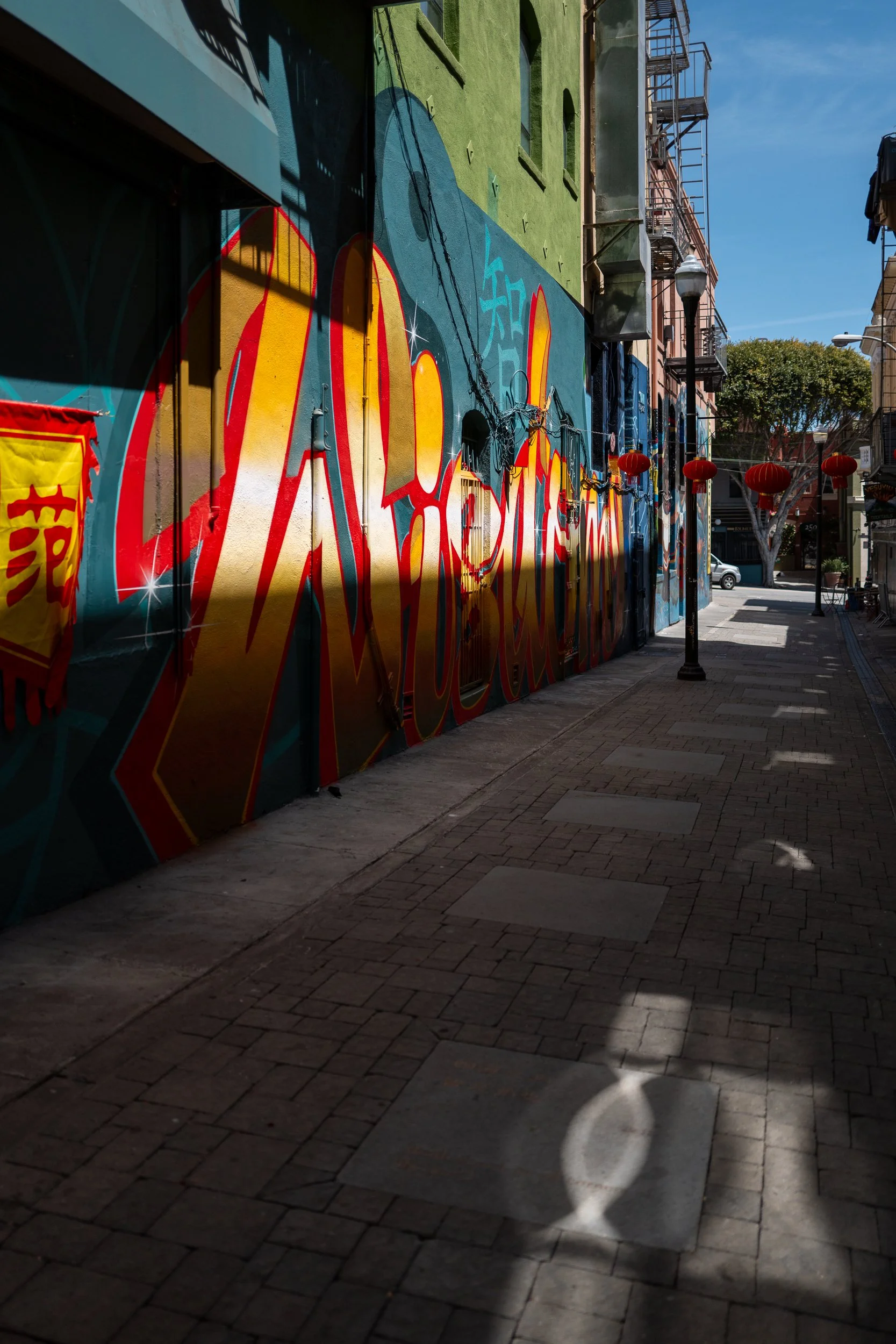 Rue piétonne avec mural de graffiti coloré, lampadaires, lanternes rouges chinoises, et ombres sur le trottoir, arbre en arrière-plan, ciel bleu.