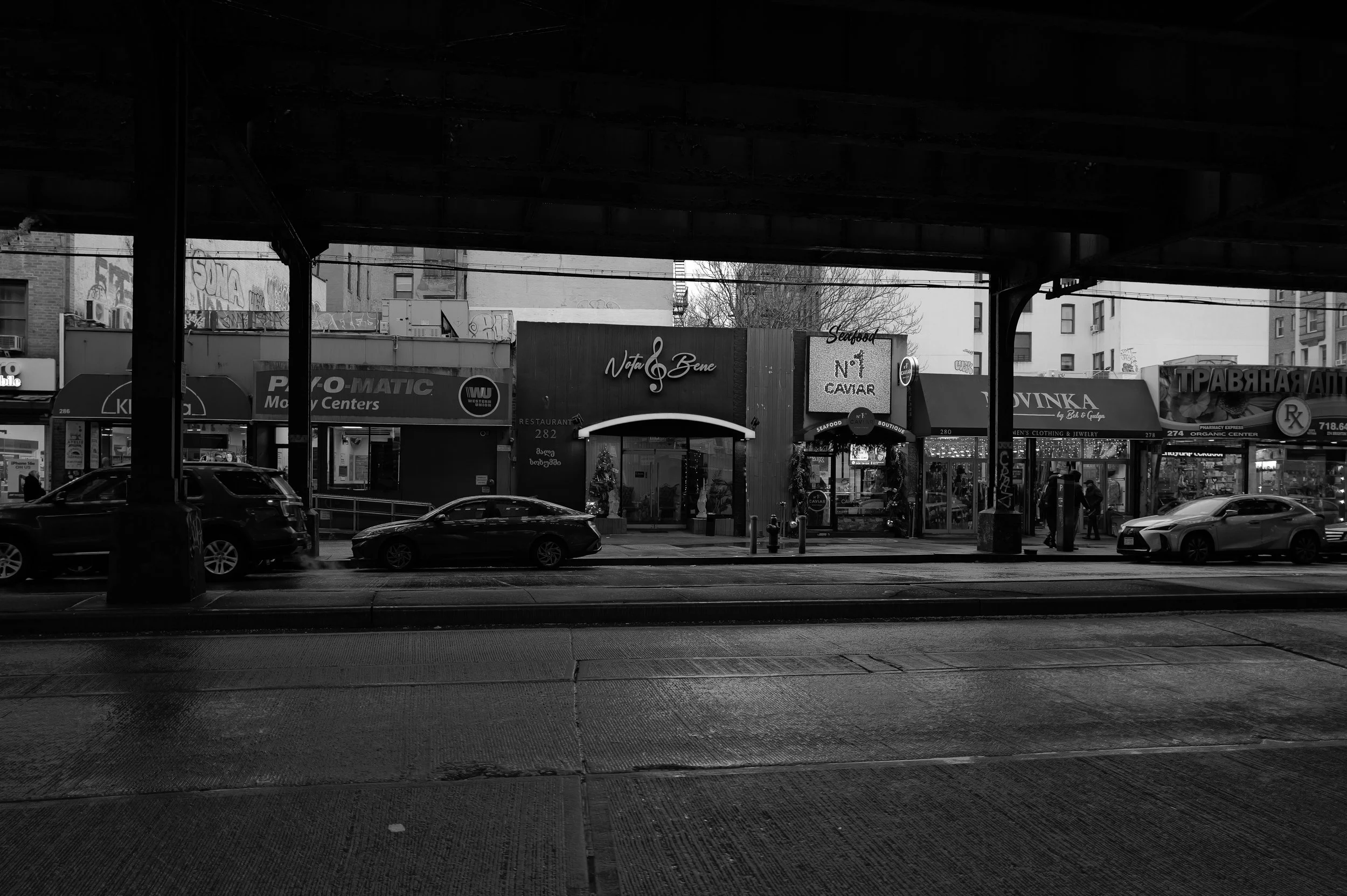 Vue depuis sous un pont sur une rue urbaine avec des voitures, commerces et enseignes, en noir et blanc.