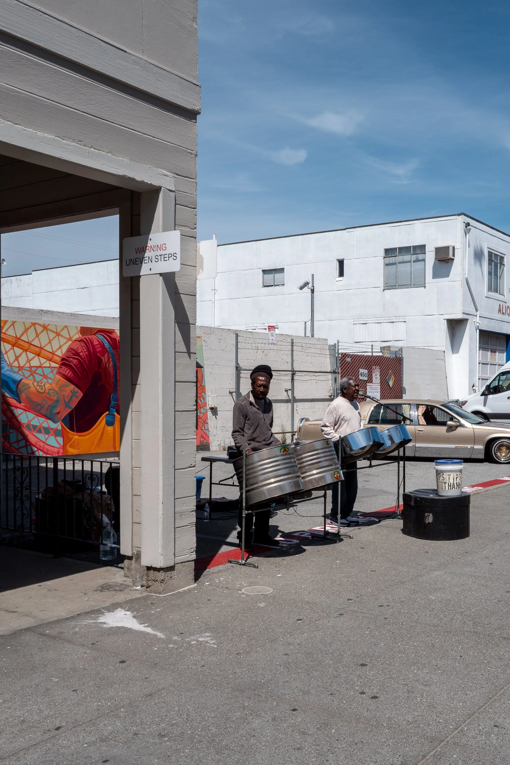 Deux hommes jouent de la steel drum dans une rue, avec des voitures et un bâtiment en arrière-plan.